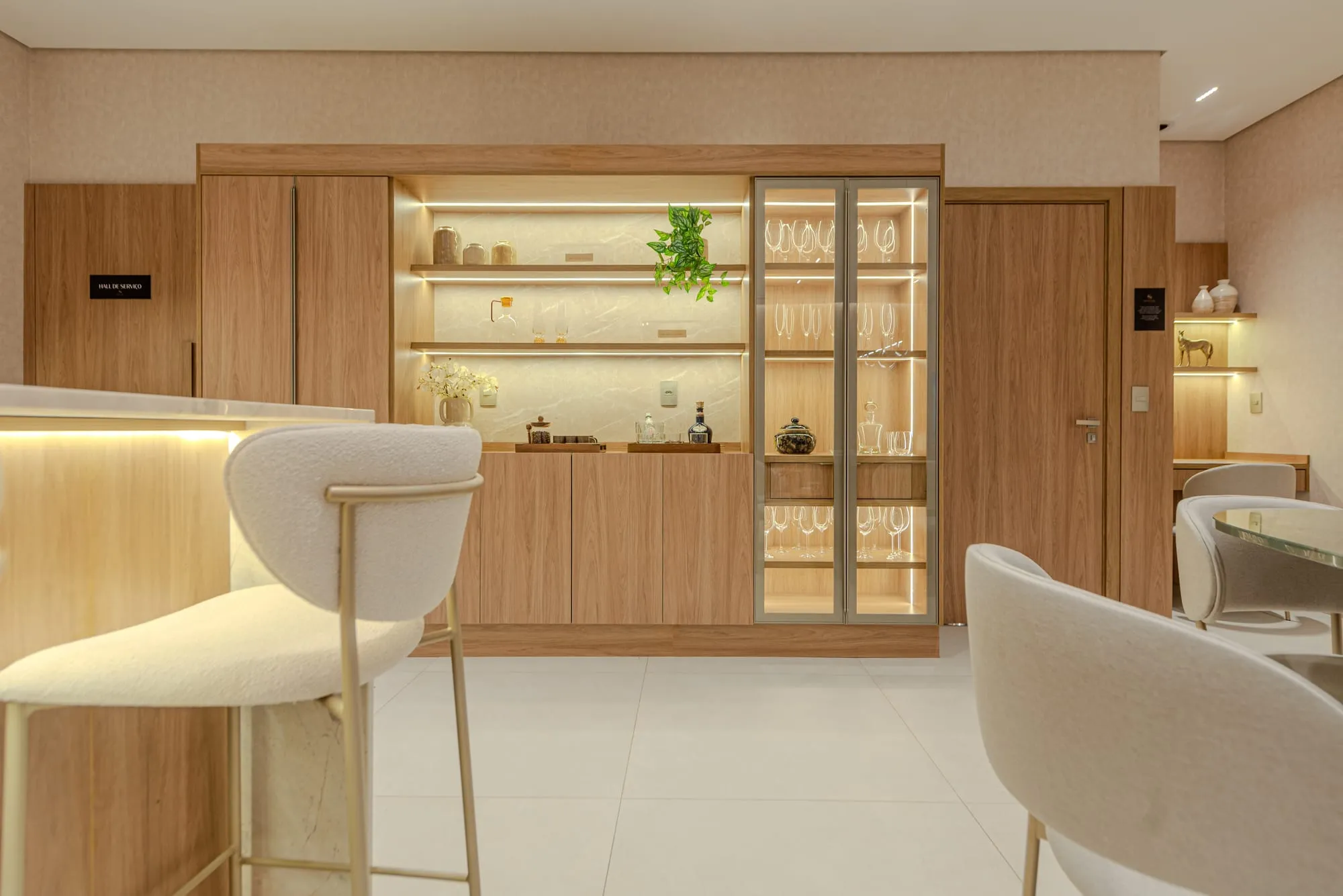 Marcenaria iluminada da cozinha com nichos, cristaleira e banqueta estofada junto à ilha