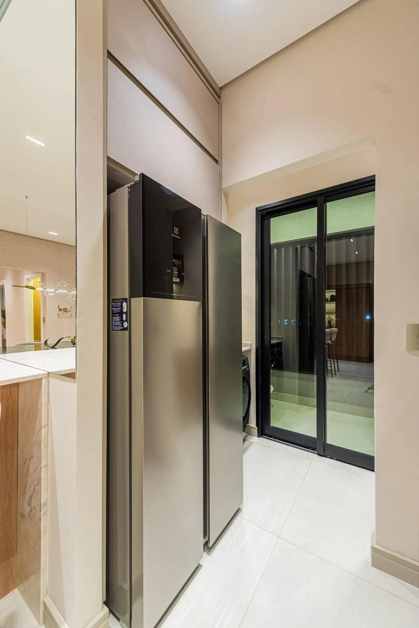 Geladeira de inox embutida e porta de vidro que leva à área de serviço com lavanderia