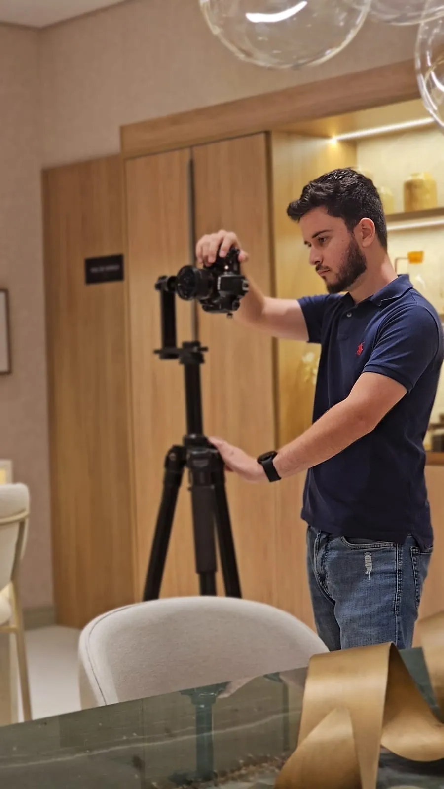 Gustavo posiciona a câmera no cabeçote panorâmico junto à cristaleira iluminada, com a mesa de jantar em primeiro plano