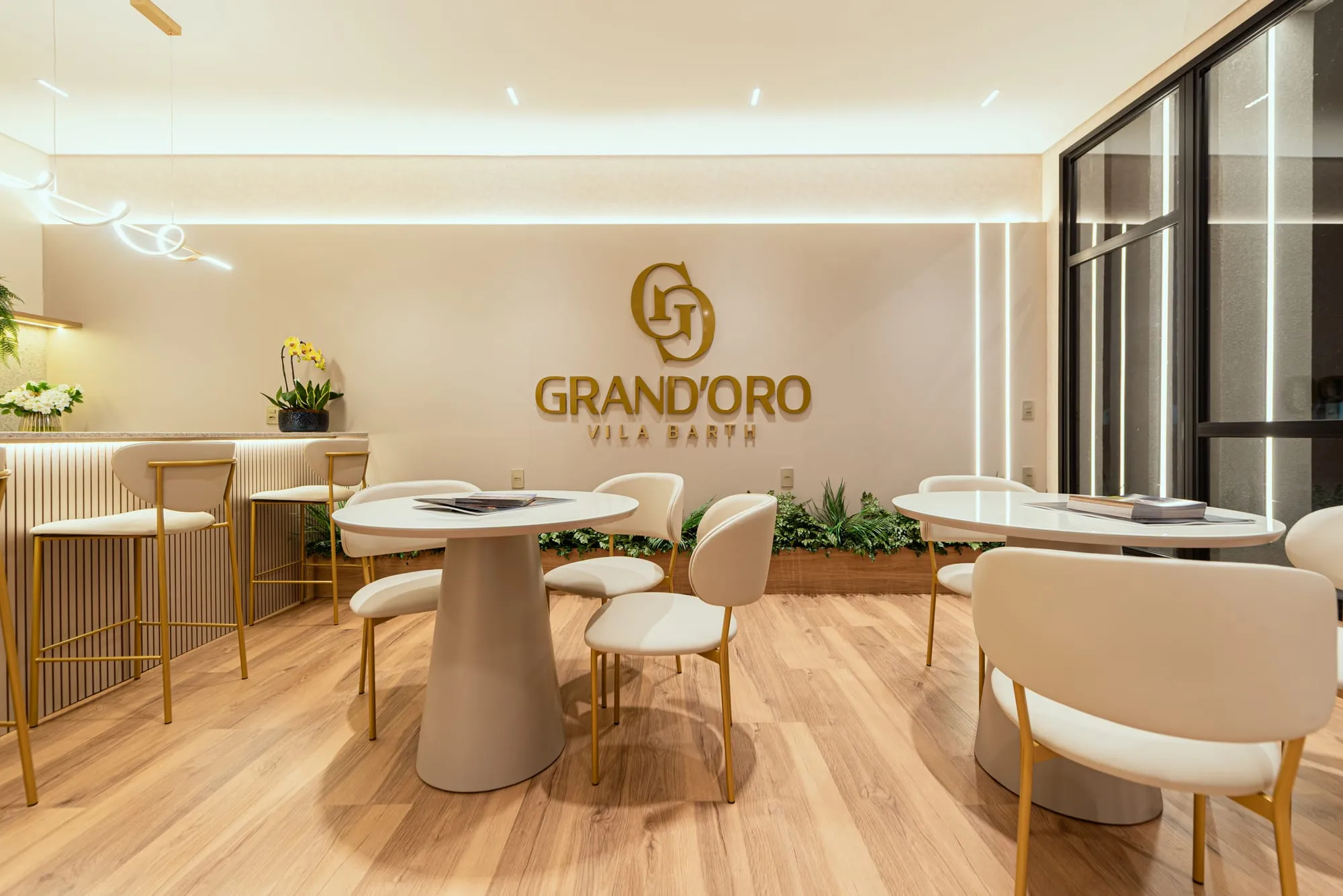 Lounge do plantão de vendas com o logo dourado do Grand'Oro Vila Barth na parede, mesas redondas e a maquete ao centro