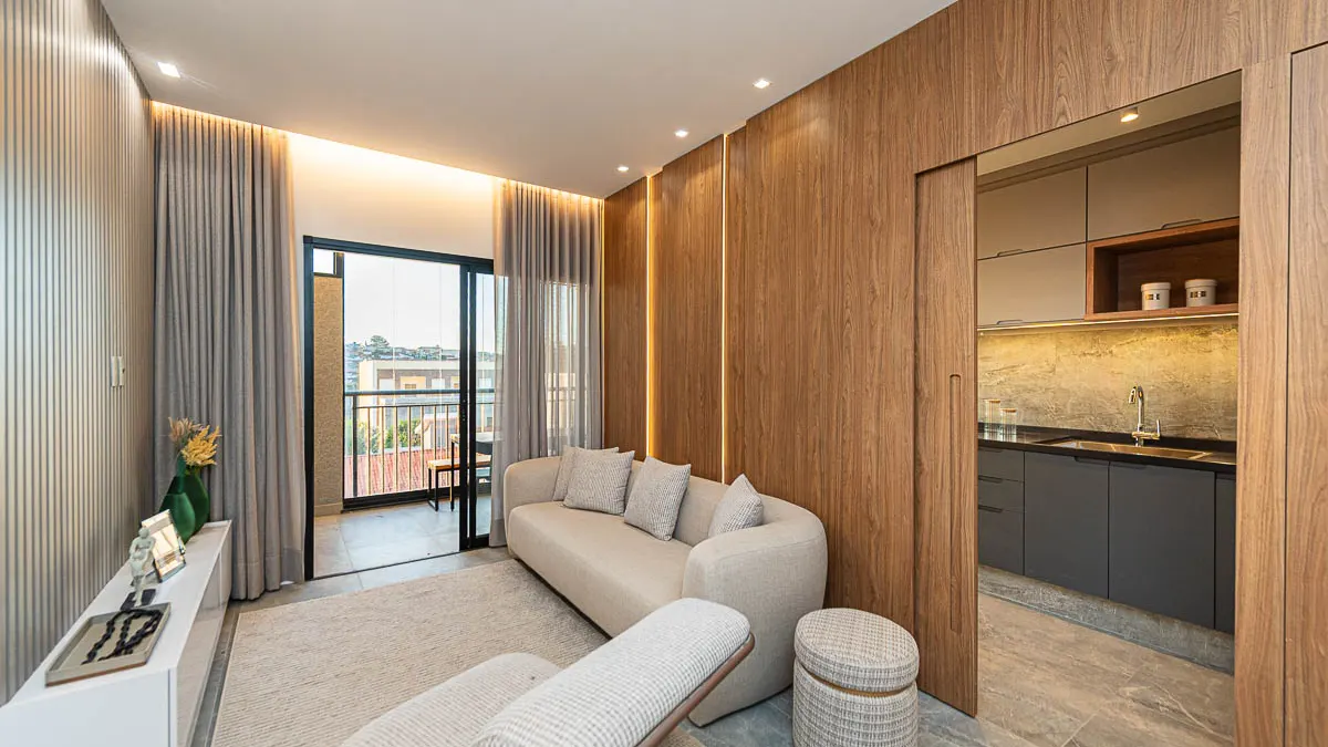 Sala de estar do decorado Haus Residence Club com sofá claro e painel de madeira, a cozinha à direita e a varanda ao fundo