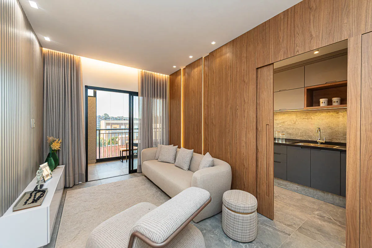 Sala de estar do decorado Haus Residence Club com sofá claro e painel de madeira, a cozinha à direita e a varanda ao fundo