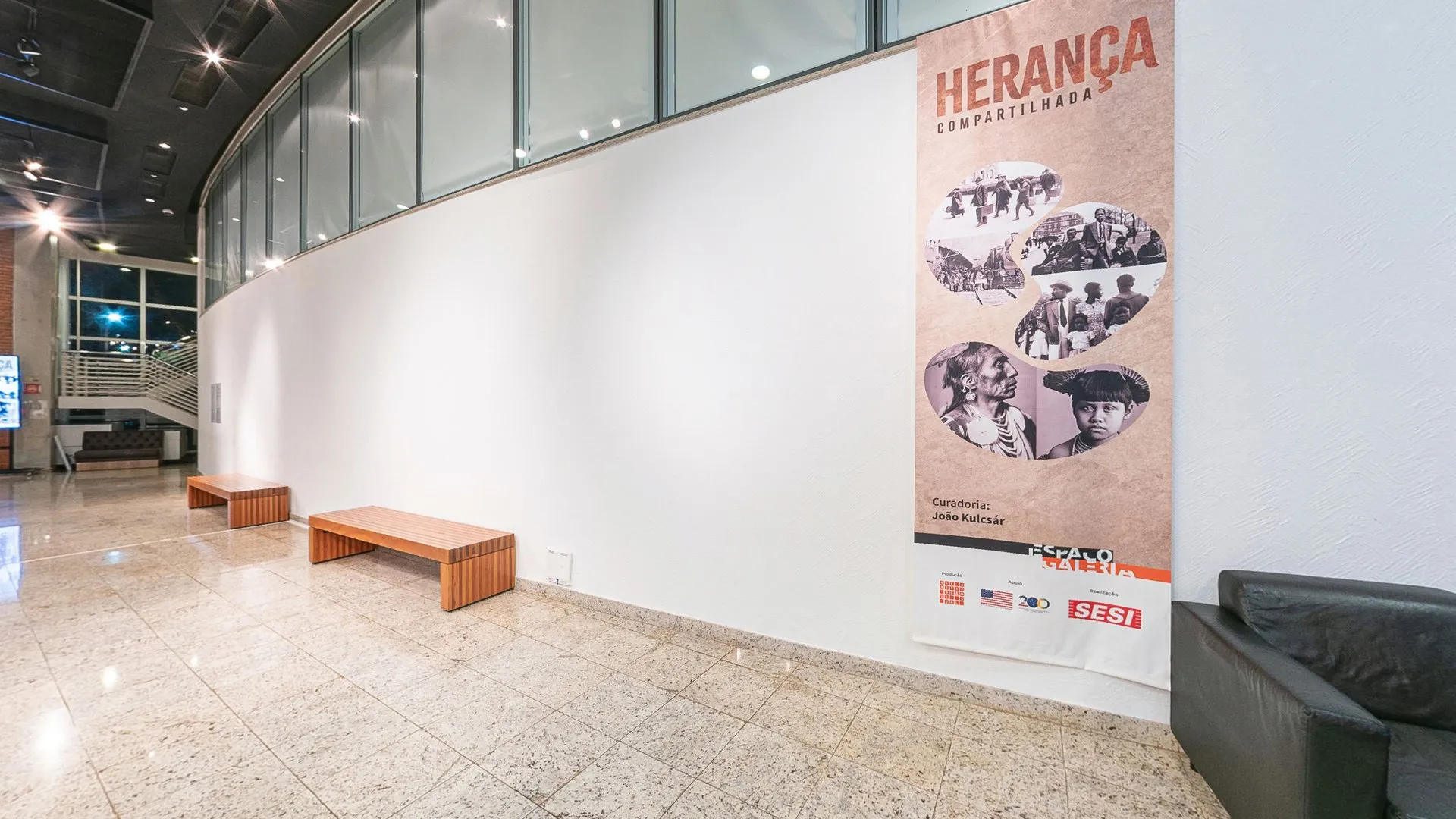 Banner da exposição Herança Compartilhada junto à parede curva do Foyer do Teatro do SESI Itapetininga, com bancos de madeira