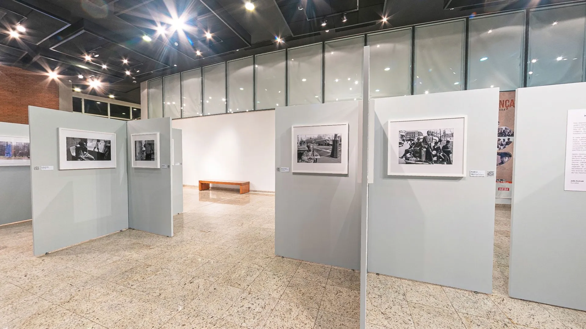 Biombos cinza da exposição Herança Compartilhada no Foyer do Teatro do SESI Itapetininga, com fotografias emolduradas e o banner da mostra ao fundo
