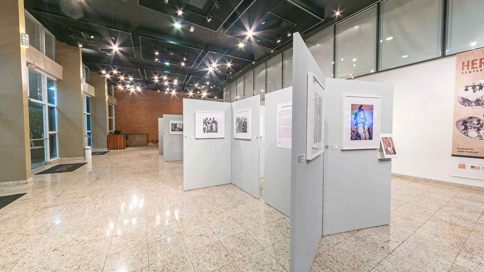 Vista ampla do Foyer do Teatro do SESI Itapetininga com os biombos da exposição Herança Compartilhada, a parede de tijolos e o banner da mostra