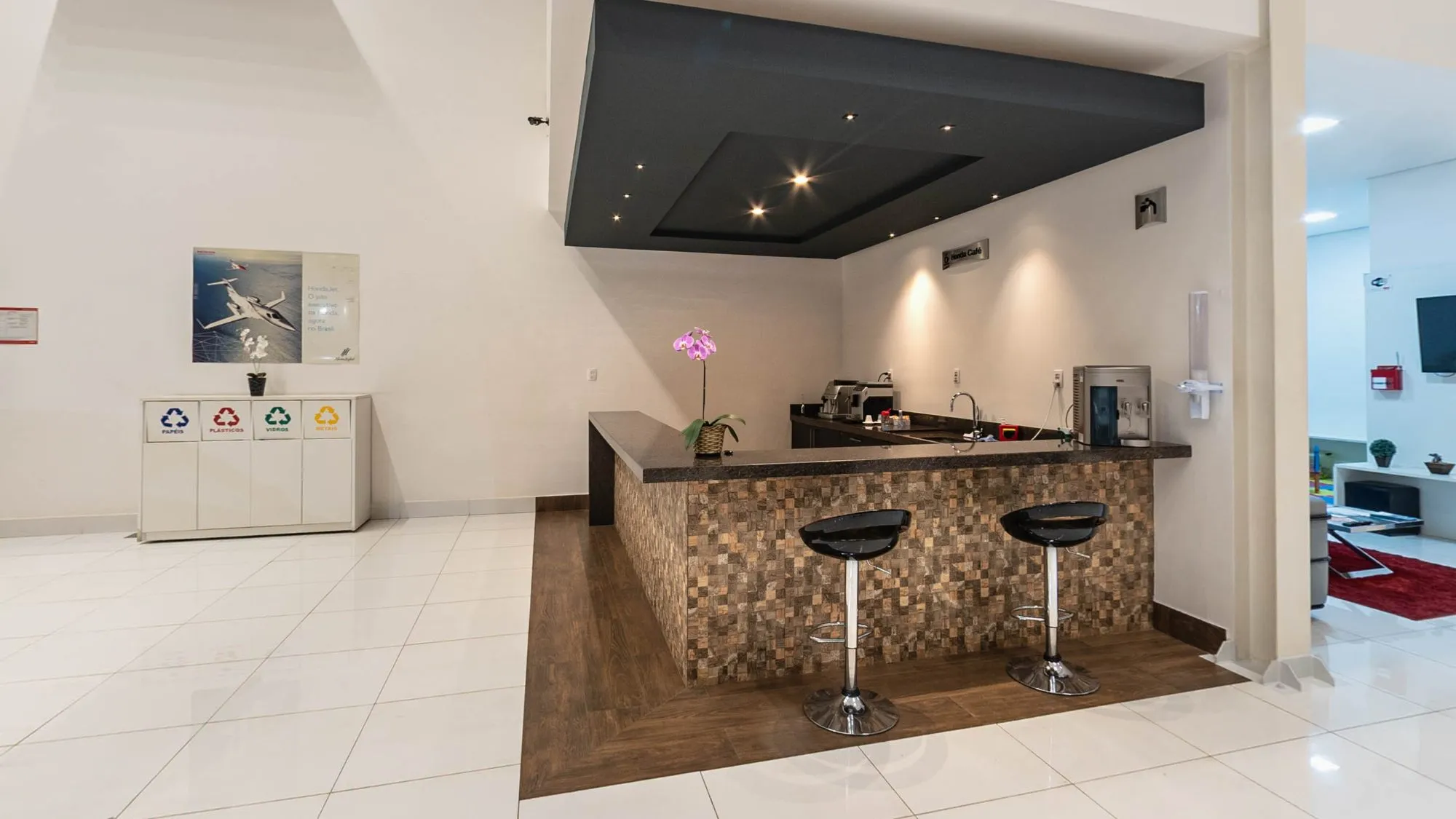 Café do showroom da Hipervel com balcão revestido de pastilhas em tons de madeira e tampo escuro, duas banquetas pretas, orquídea e máquina de café, lixeiras de coleta seletiva à esquerda e a sala de espera com tapete vermelho à direita