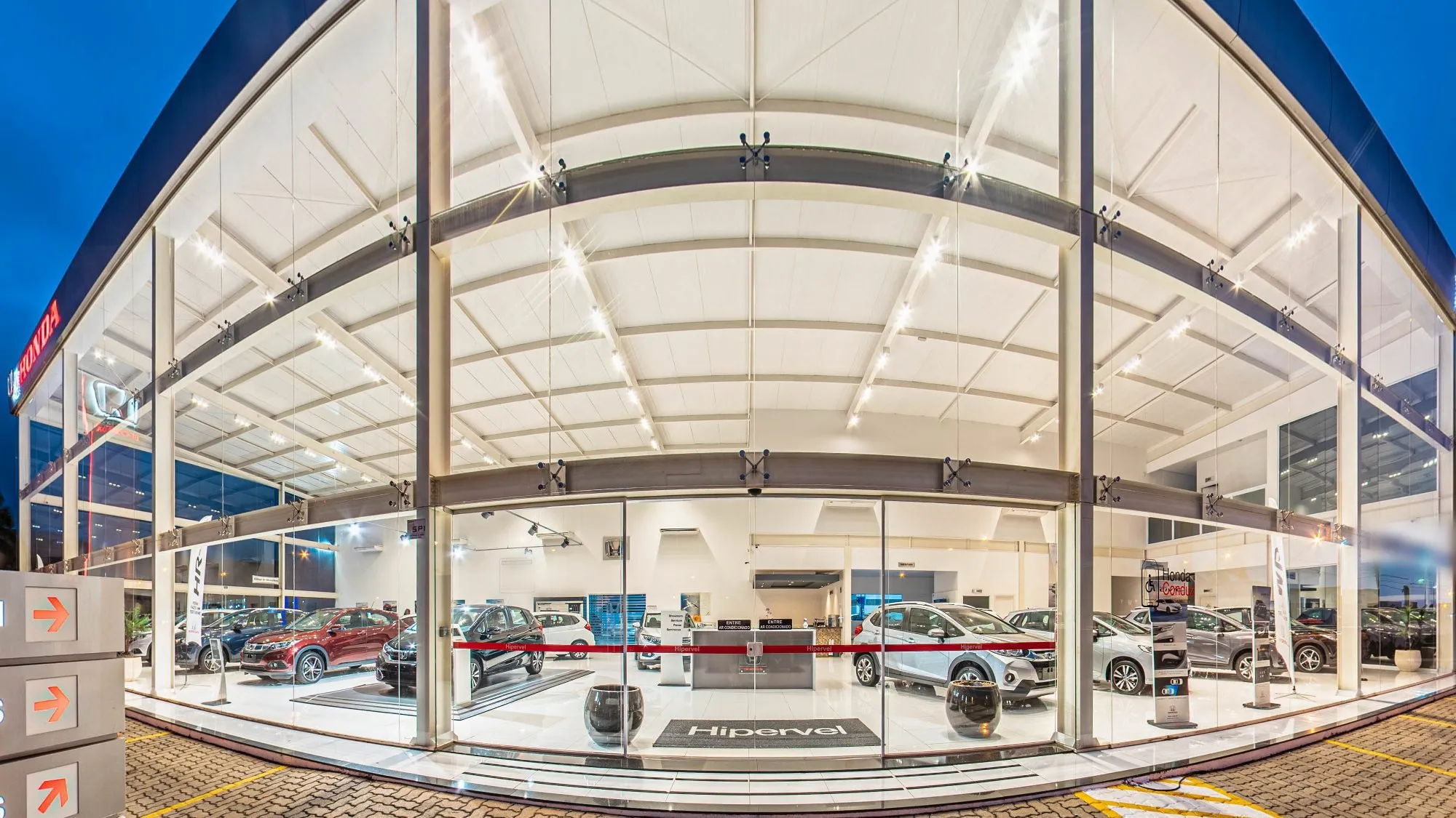 Fachada de vidro da Honda Hipervel em Itapetininga ao anoitecer, com a cobertura curva de vigas brancas iluminada por dentro, carros expostos atrás do vidro, o tapete com a marca Hipervel na entrada e o letreiro vermelho da Honda à esquerda
