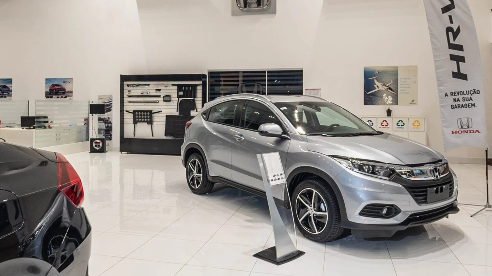Honda HR-V prata de três quartos de frente no showroom da Hipervel, ao lado de um totem de informações, com a bandeira HR-V e a parede de acessórios ao fundo e a lateral traseira de um Civic preto no canto