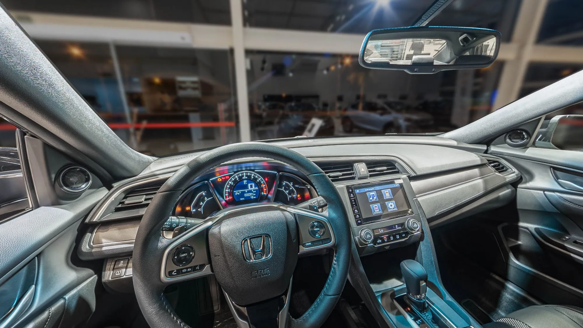 Interior do Honda Civic visto do banco do motorista: volante com o logo Honda, painel digital aceso, central multimídia, alavanca do câmbio e o showroom desfocado pelo para-brisa
