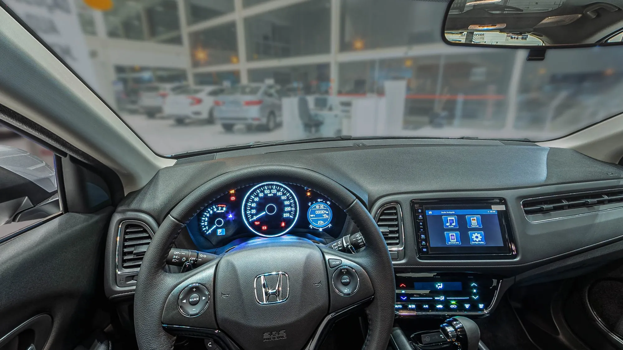 Interior do Honda HR-V visto do banco do motorista: volante com o logo Honda, painel com três mostradores iluminados em azul, central multimídia, comandos do ar-condicionado e o showroom desfocado pelo para-brisa