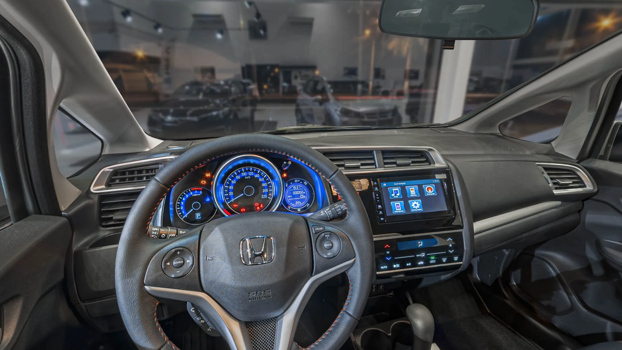 Interior do Honda WR-V visto do banco do motorista: volante com costura laranja e o logo Honda, painel com mostradores em azul, central multimídia e o showroom escuro pelo para-brisa