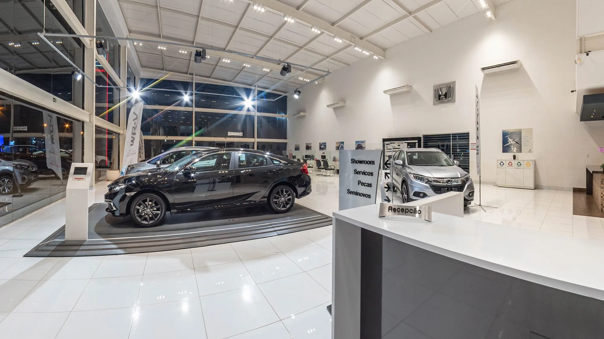 Recepção da Hipervel com o balcão e a placa Recepção em primeiro plano, a placa de direção para Showroom, Serviços, Peças e Seminovos, um Honda Civic preto sobre pedestal escuro à esquerda e um HR-V prata ao fundo