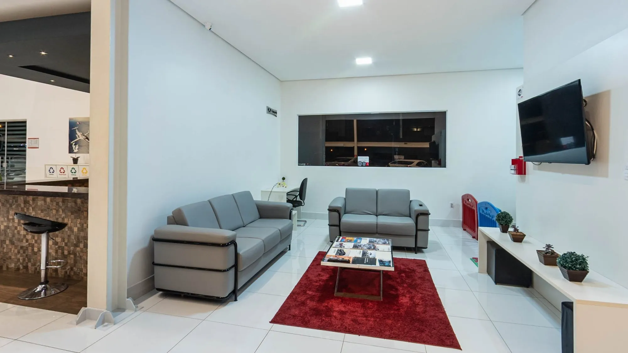 Sala de espera da Hipervel com dois sofás cinza, mesa de centro com revistas sobre tapete vermelho, TV na parede, janela ao fundo e prateleira baixa com vasos