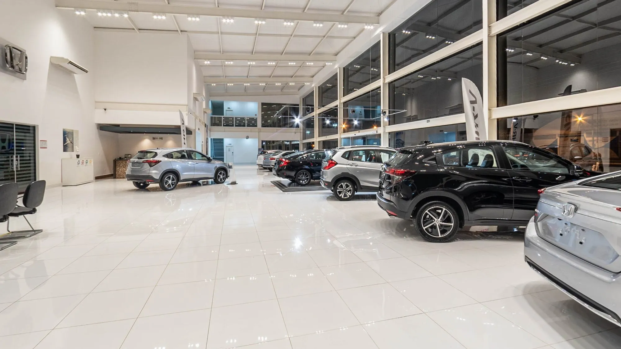 Salão do showroom da Honda Hipervel em Itapetininga no tour 360°, com piso branco brilhante, pé-direito alto e fachada de vidro, um HR-V prata no meio, um carro preto sobre pedestal ao fundo, um WR-V prata e outros carros enfileirados