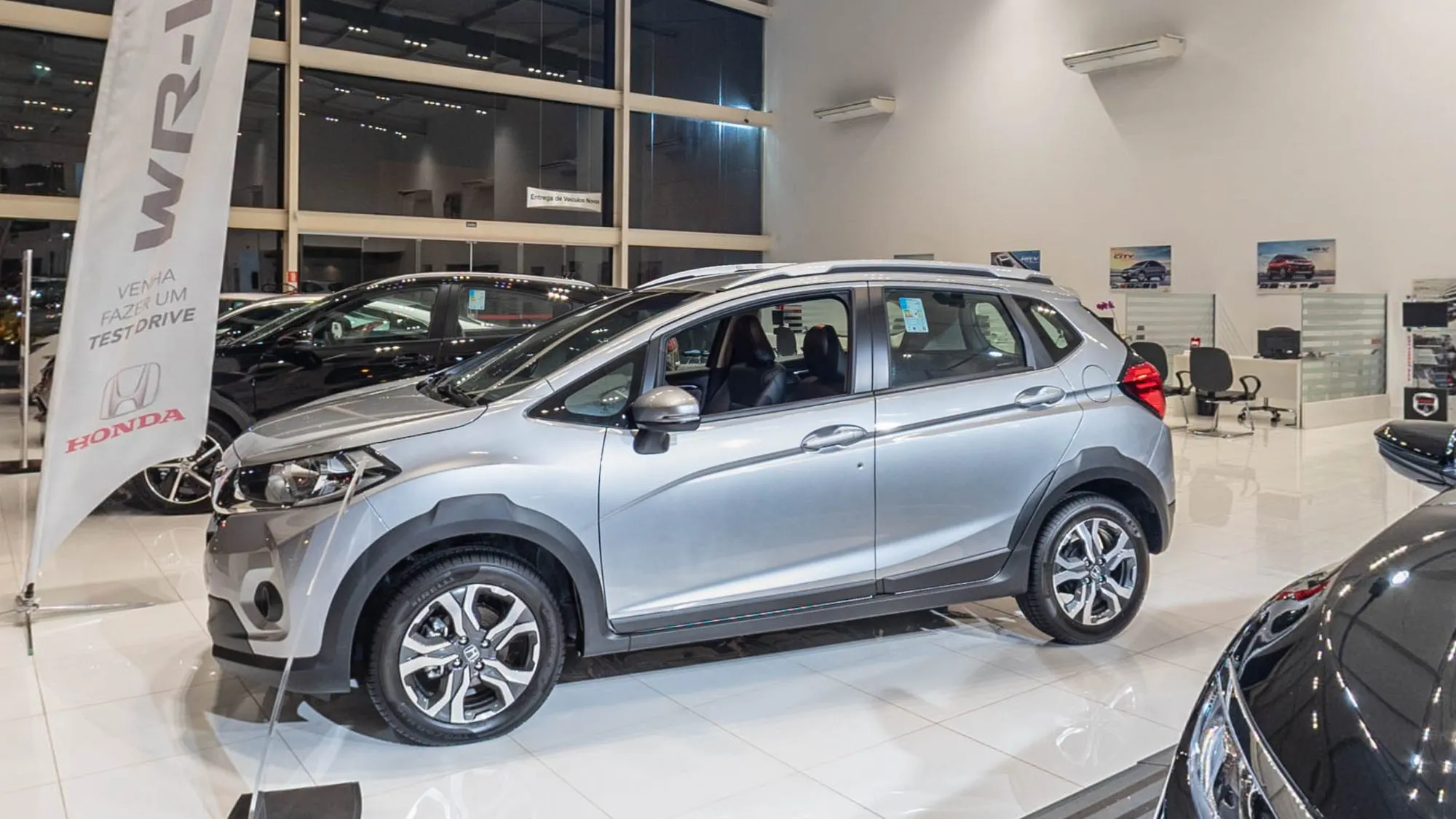 Honda WR-V prata de perfil no showroom da Hipervel, com rack de teto, ao lado de uma bandeira WR-V da Honda, com a fachada de vidro, carros pretos e as mesas de atendimento ao fundo