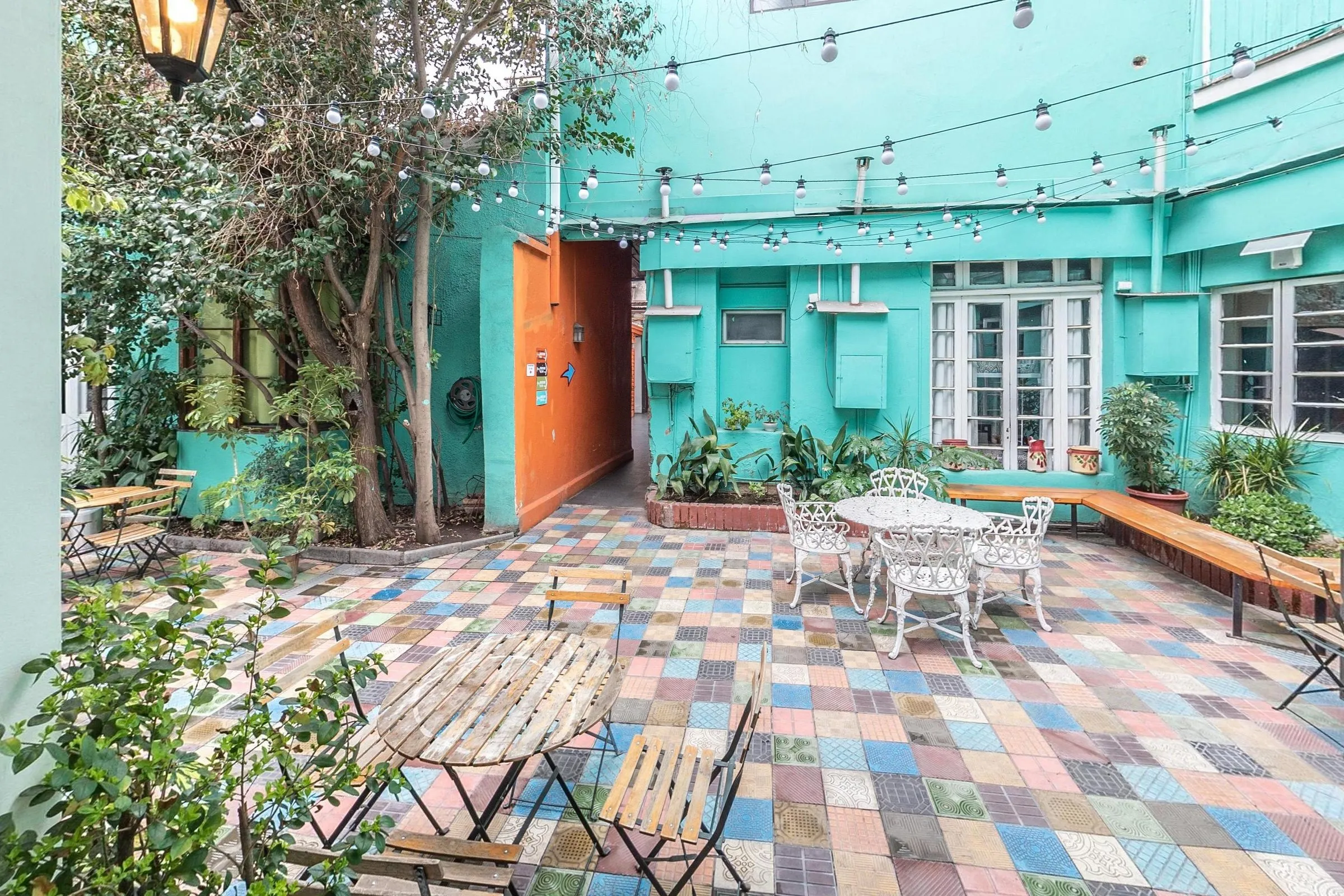 Pátio interno do hostal com piso de ladrilhos coloridos, mesa redonda e cadeiras de ferro fundido branco, mesa dobrável de ripas de madeira, banco de madeira junto ao canteiro, varais de lâmpadas e paredes verde-água com janelas de caixilho branco e uma passagem laranja