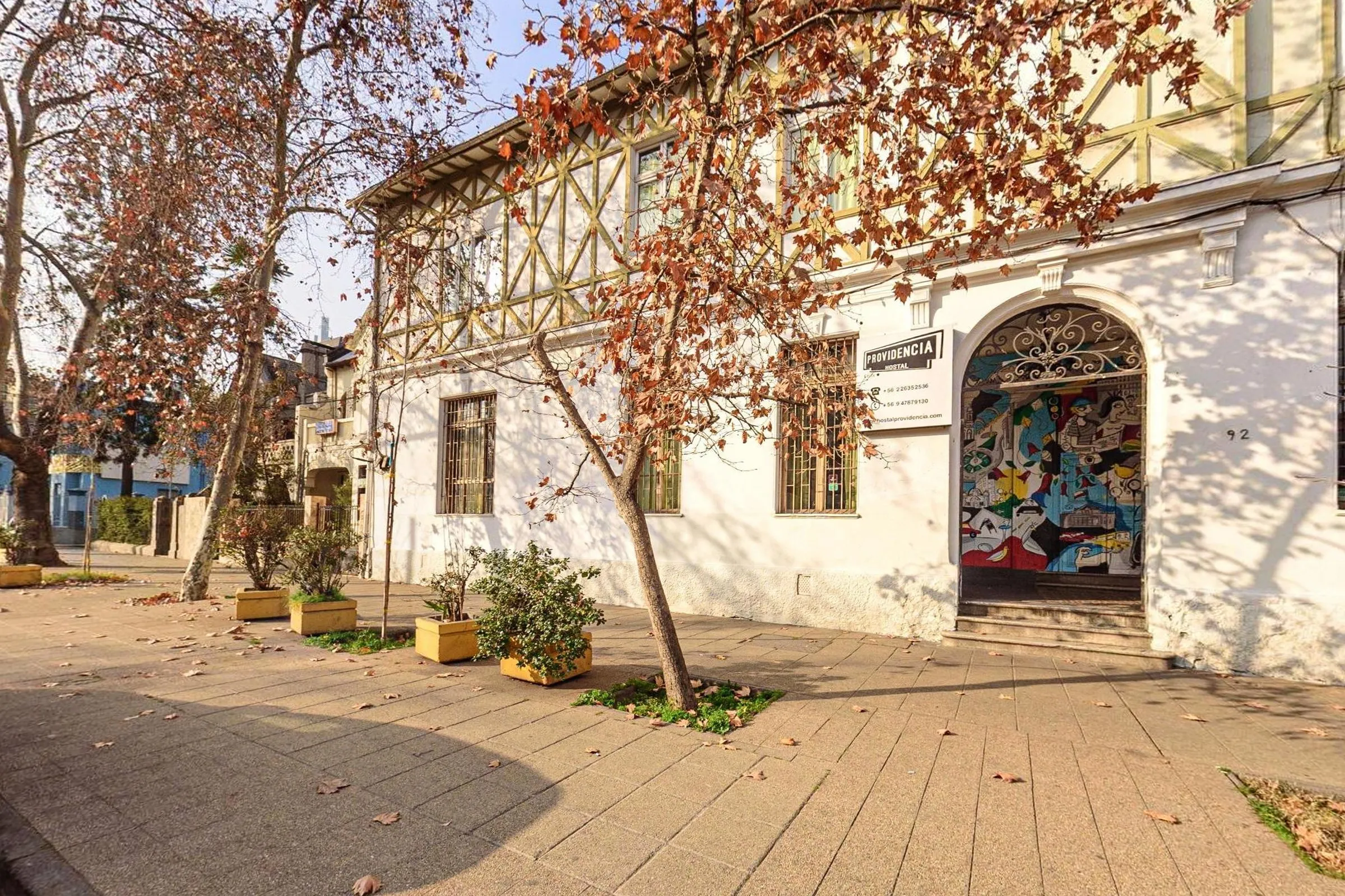 Fachada de esquina do Hostal Providencia, em Santiago, um casarão de paredes brancas com enxaimel no andar de cima, janelas de madeira e a entrada em arco com mural colorido, atrás de uma árvore de folhas secas e floreiras na calçada