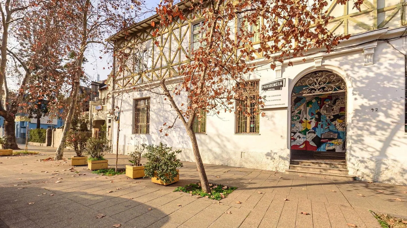 Fachada de esquina do Hostal Providencia, em Santiago, um casarão de paredes brancas com enxaimel no andar de cima, janelas de madeira e a entrada em arco com mural colorido, atrás de uma árvore de folhas secas e floreiras na calçada