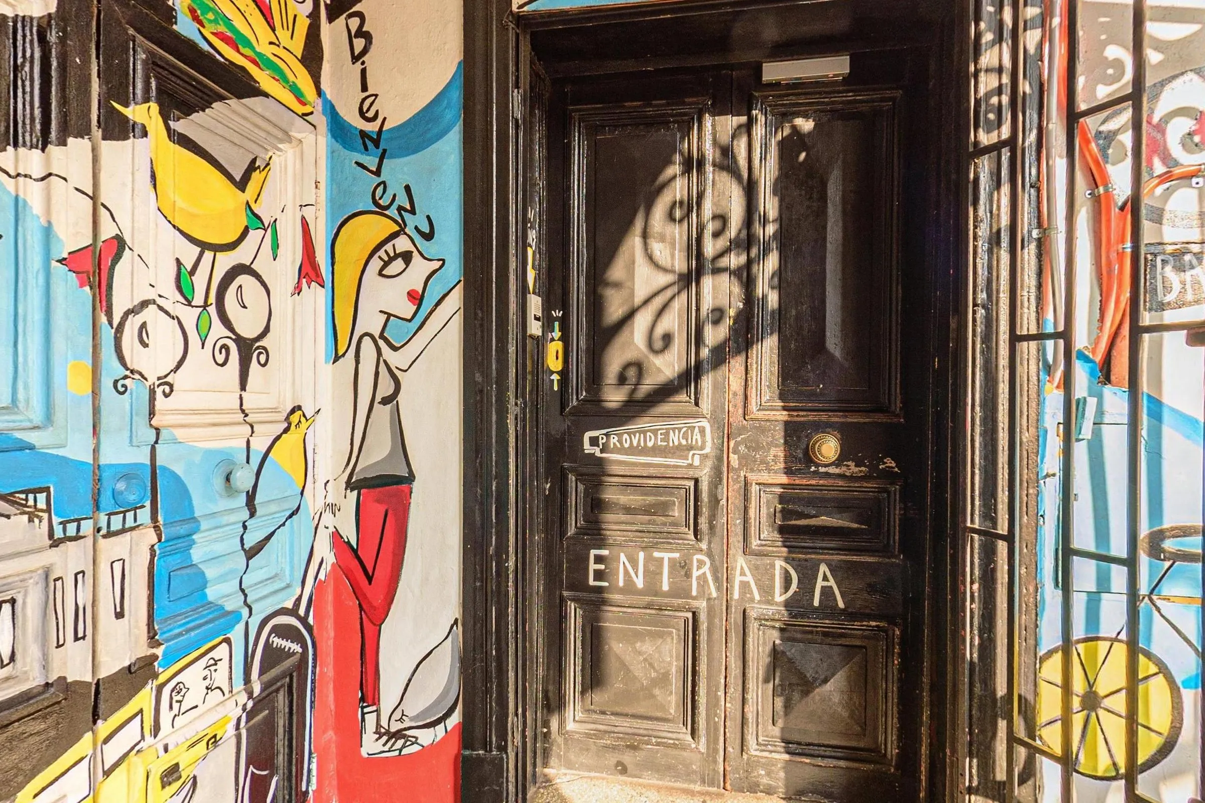 Porta dupla antiga de madeira escura da entrada do Hostal Providencia, com ENTRADA escrito à mão e a placa Providencia, entre paredes do vestíbulo pintadas com mural colorido de figuras, pássaros e prédios