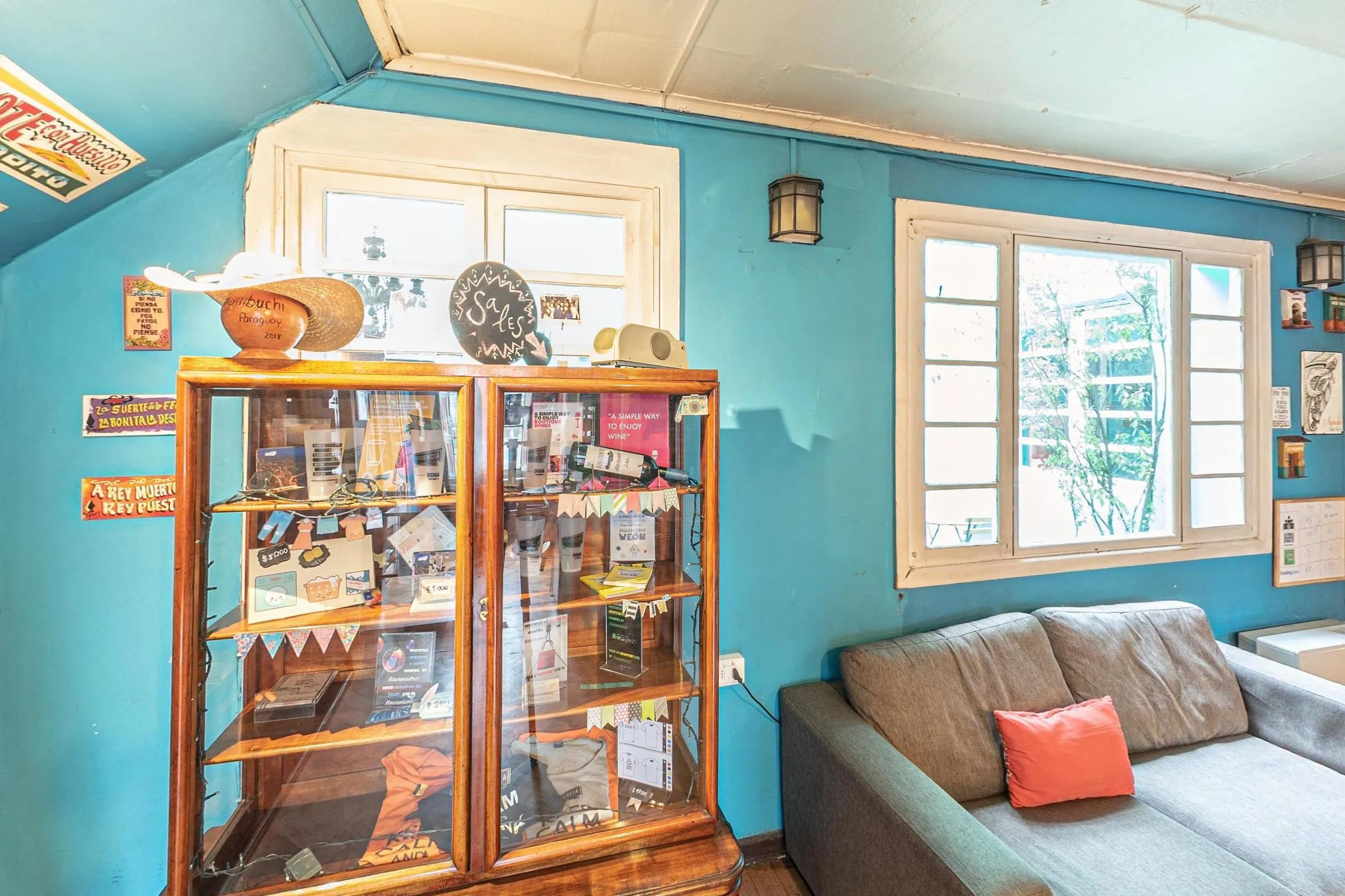 Lounge da recepção do Hostal Providencia, com paredes azuis, cristaleira antiga de madeira e vidro com objetos e lembranças, sofá cinza com almofada coral, janelas de caixilho branco e piso de taco em xadrez