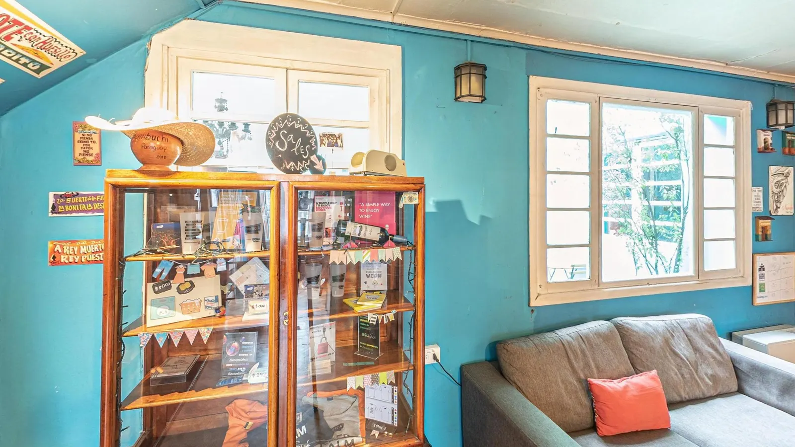 Lounge da recepção do Hostal Providencia, com paredes azuis, cristaleira antiga de madeira e vidro com objetos e lembranças, sofá cinza com almofada coral, janelas de caixilho branco e piso de taco em xadrez