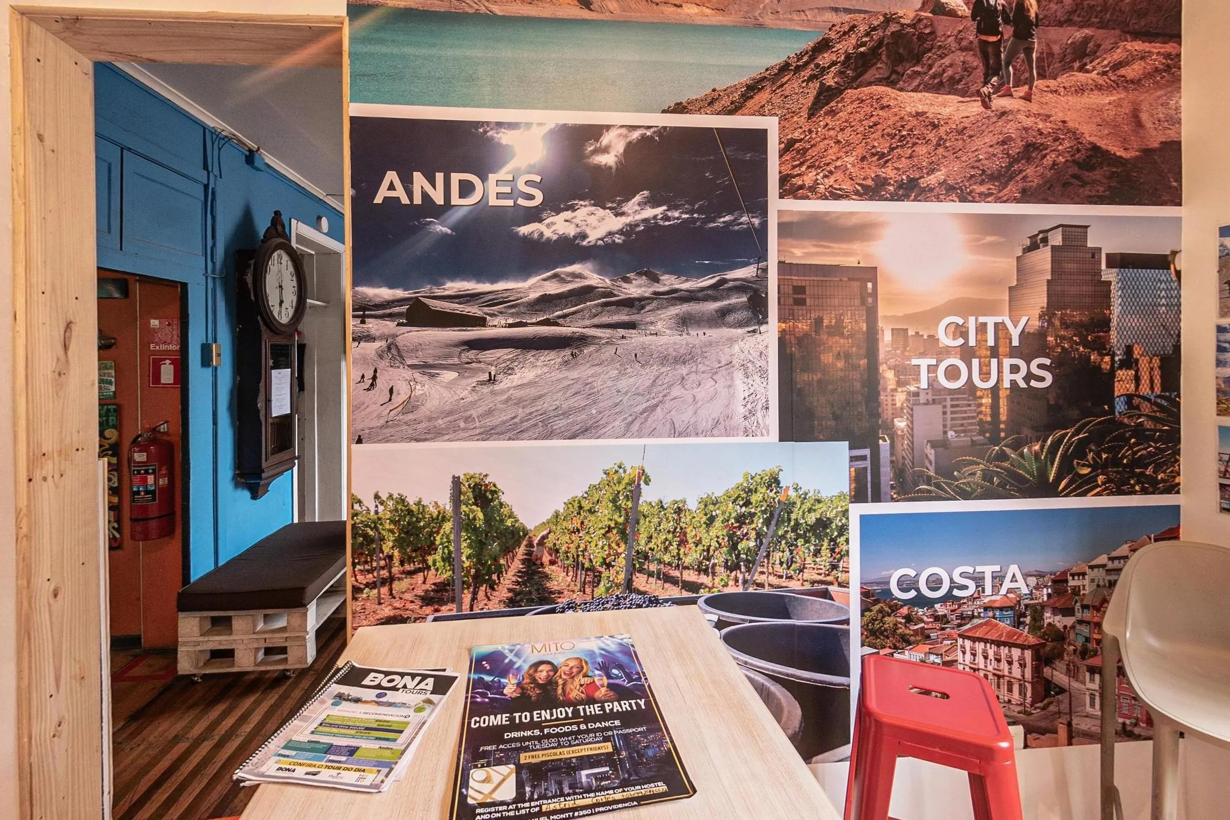 Balcão de tours com mesa de madeira clara, folhetos, banqueta vermelha e painel de fotos de passeios com os títulos Andes, City Tours e Costa; à esquerda, um relógio de parede e um sofá de pallet
