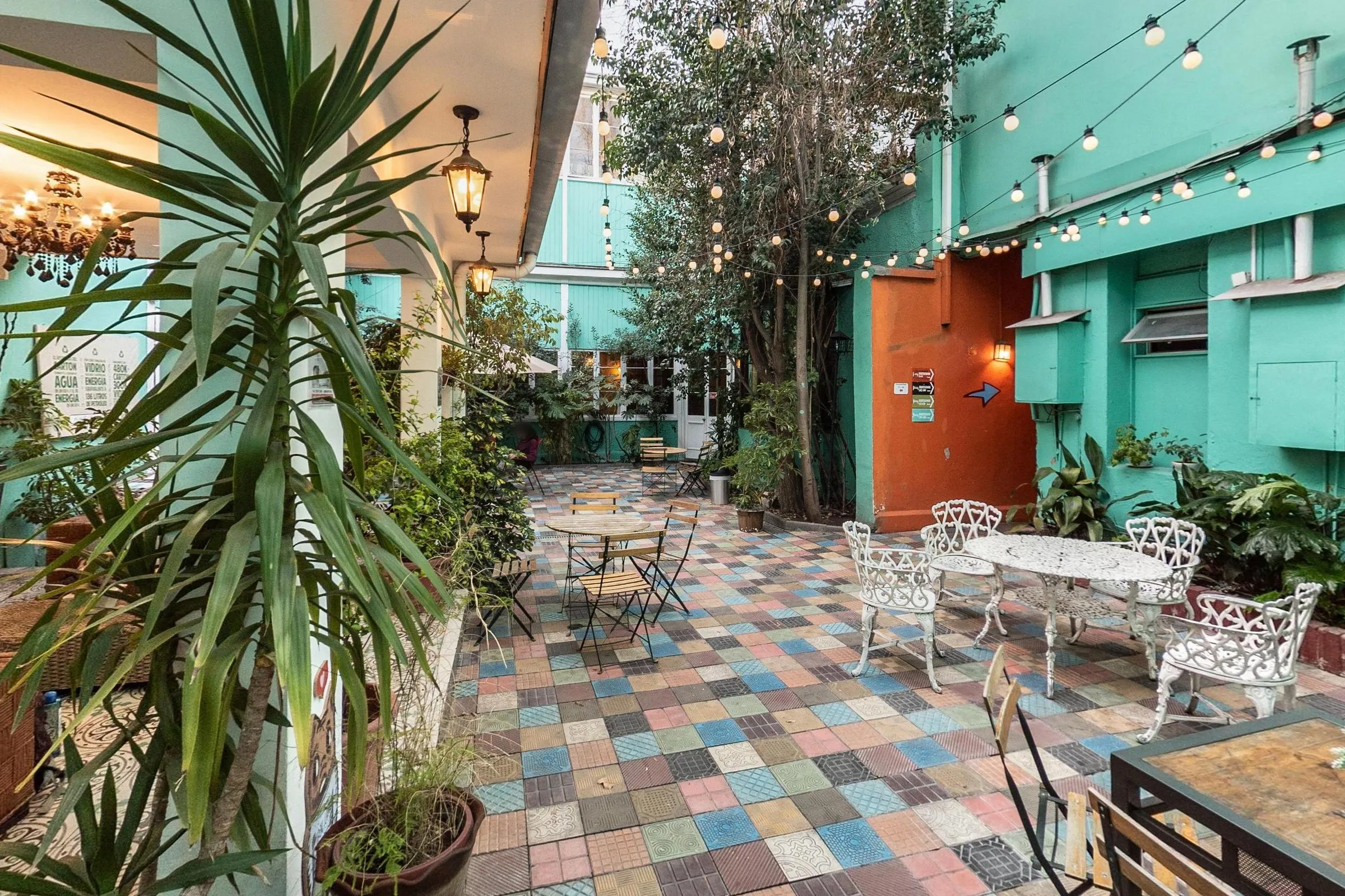 Pátio de ladrilhos coloridos visto do lounge coberto, com uma palmeira em vaso no primeiro plano, mesas e cadeiras de ferro branco, mesas dobráveis de madeira, árvores, varais de lâmpadas e paredes verde-água com uma porta laranja