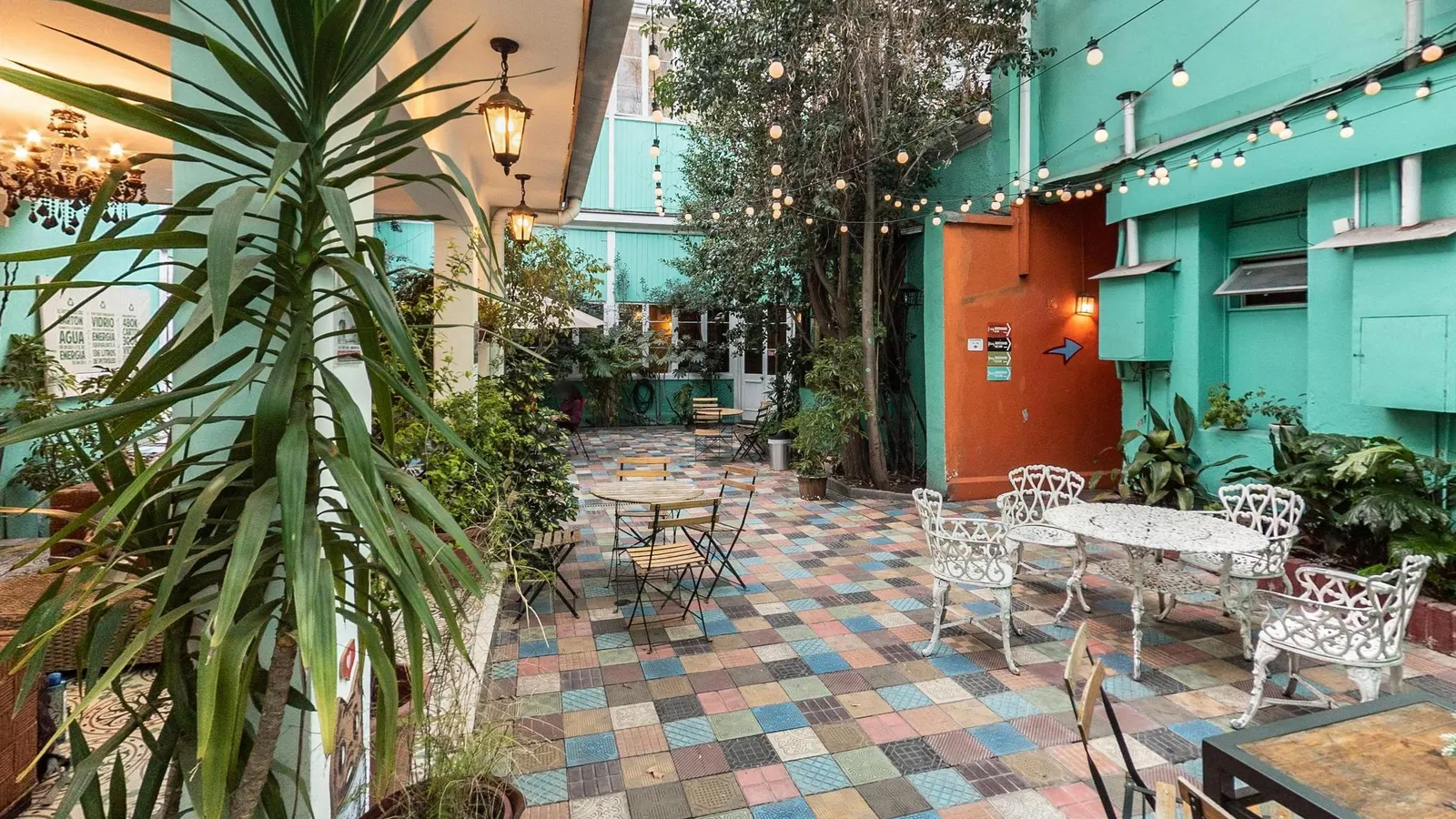 Hostal Providencia patio seen from the covered lounge, with a potted palm in the foreground, white iron tables and chairs, folding wooden tables, trees, string lights and turquoise walls with an orange door