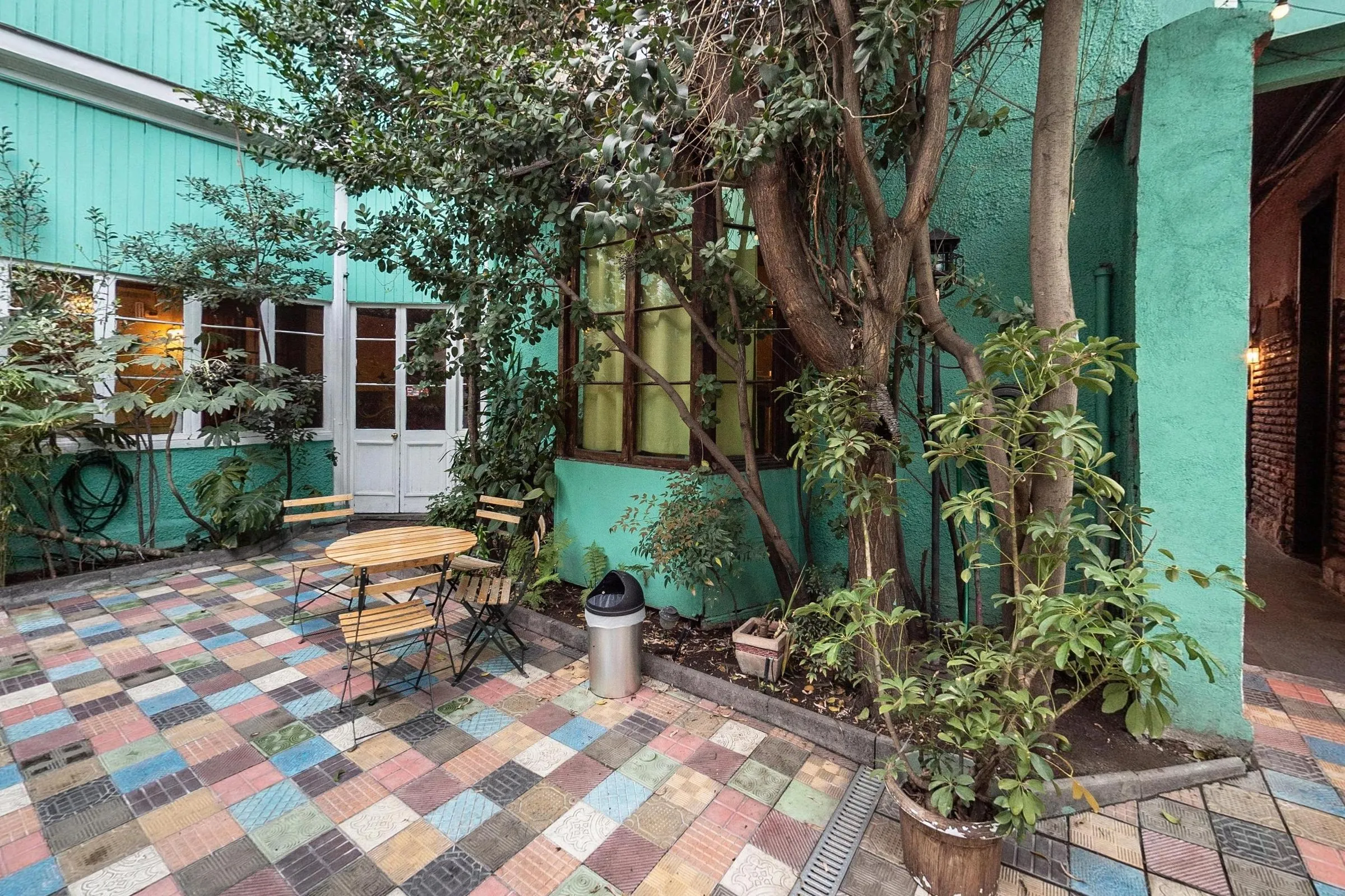 Canto ajardinado do pátio com uma árvore de tronco grosso, canteiro de plantas, mesinhas e cadeiras dobráveis de madeira sobre ladrilhos coloridos e paredes verde-água com portas brancas envidraçadas