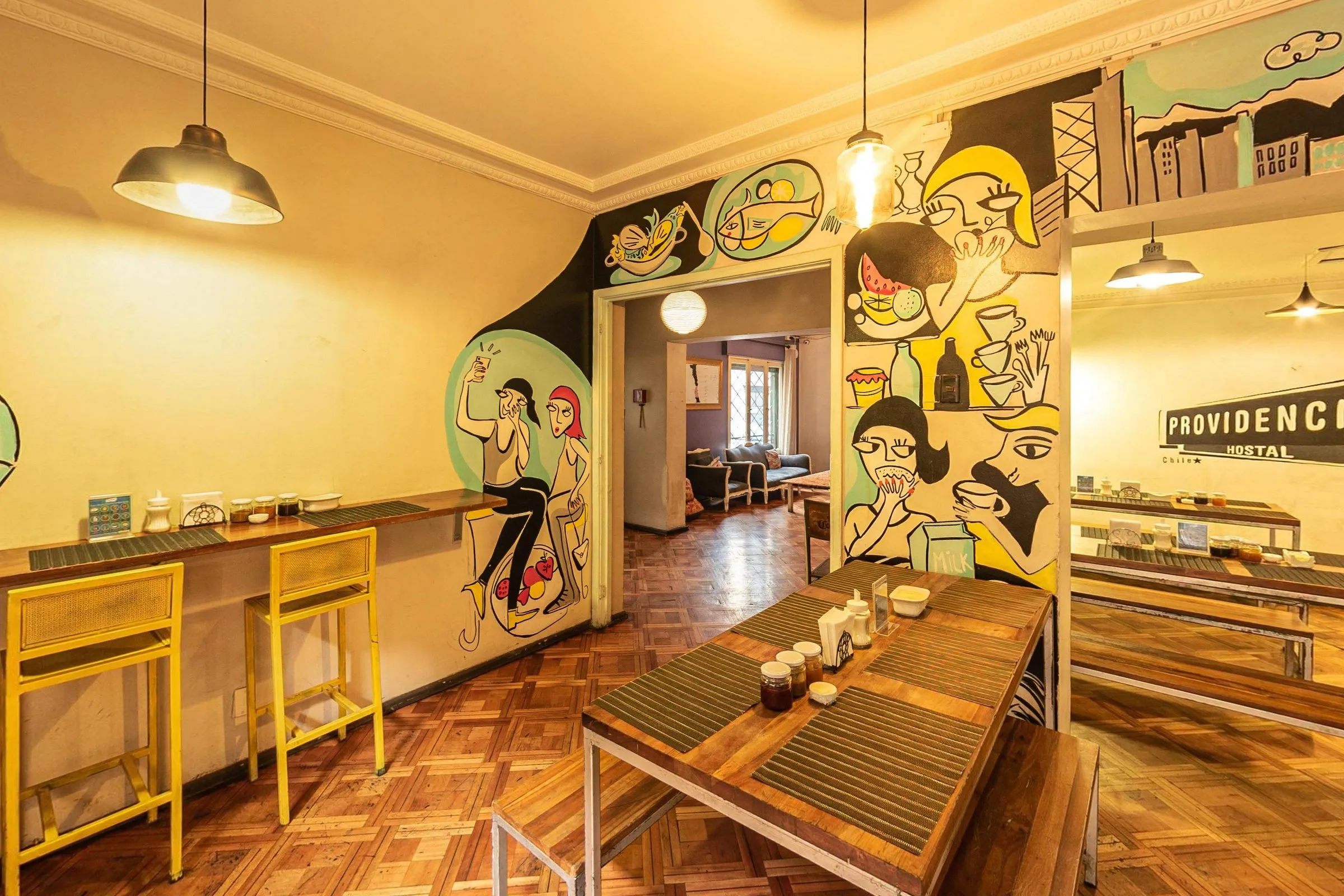Refeitório com piso de taco, mesas compridas de madeira com bancos, bancada de parede com banquetas amarelas, luminárias pendentes e mural de figuras desenhadas em preto, amarelo e verde-água; ao fundo, a placa Providencia Hostal