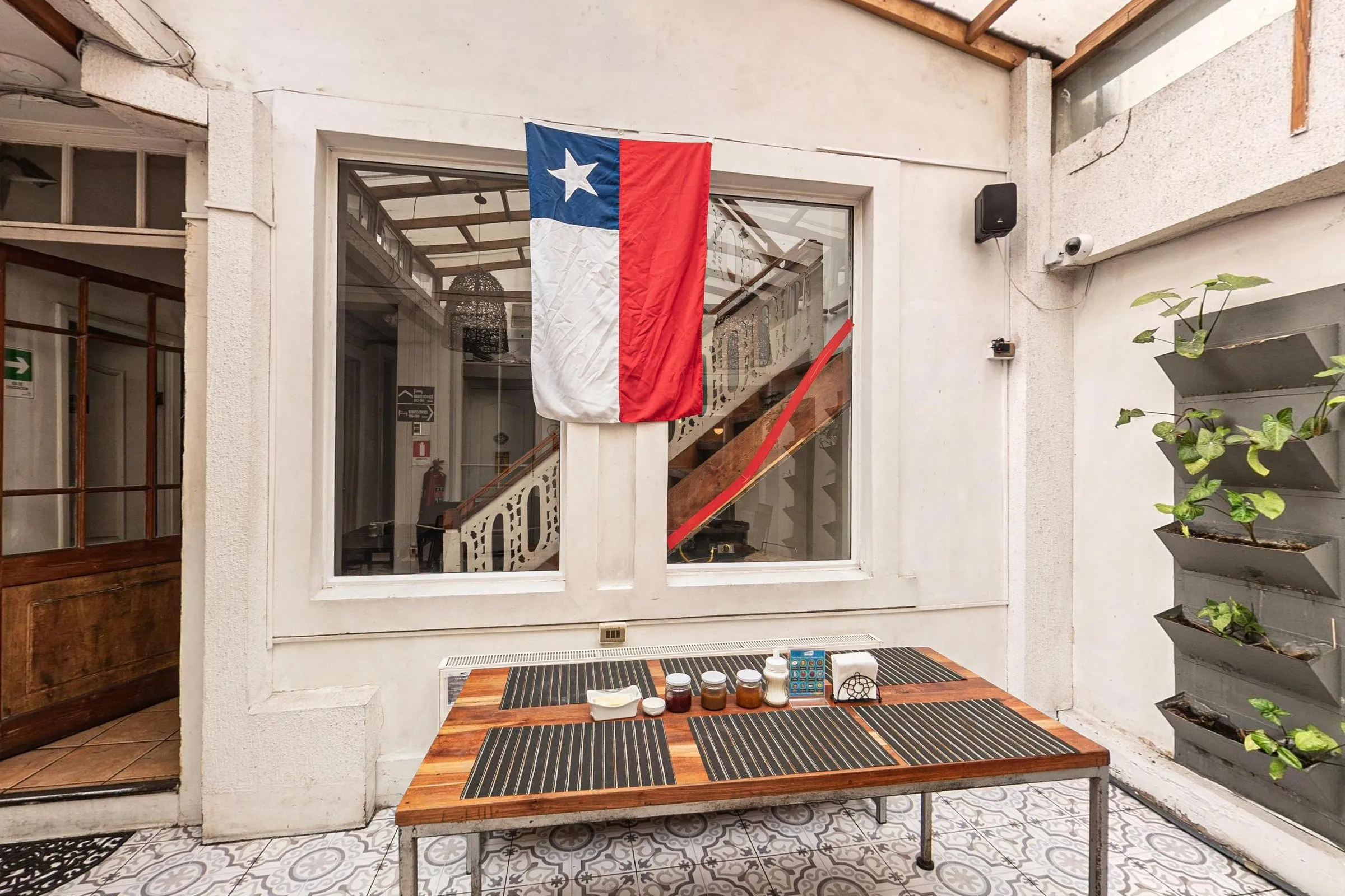 Salão do café da manhã com paredes brancas e cobertura translúcida, mesa de madeira com jogos americanos e potes de geleia, jardim vertical de vasos e a bandeira do Chile pendurada numa janela que dá para a escada
