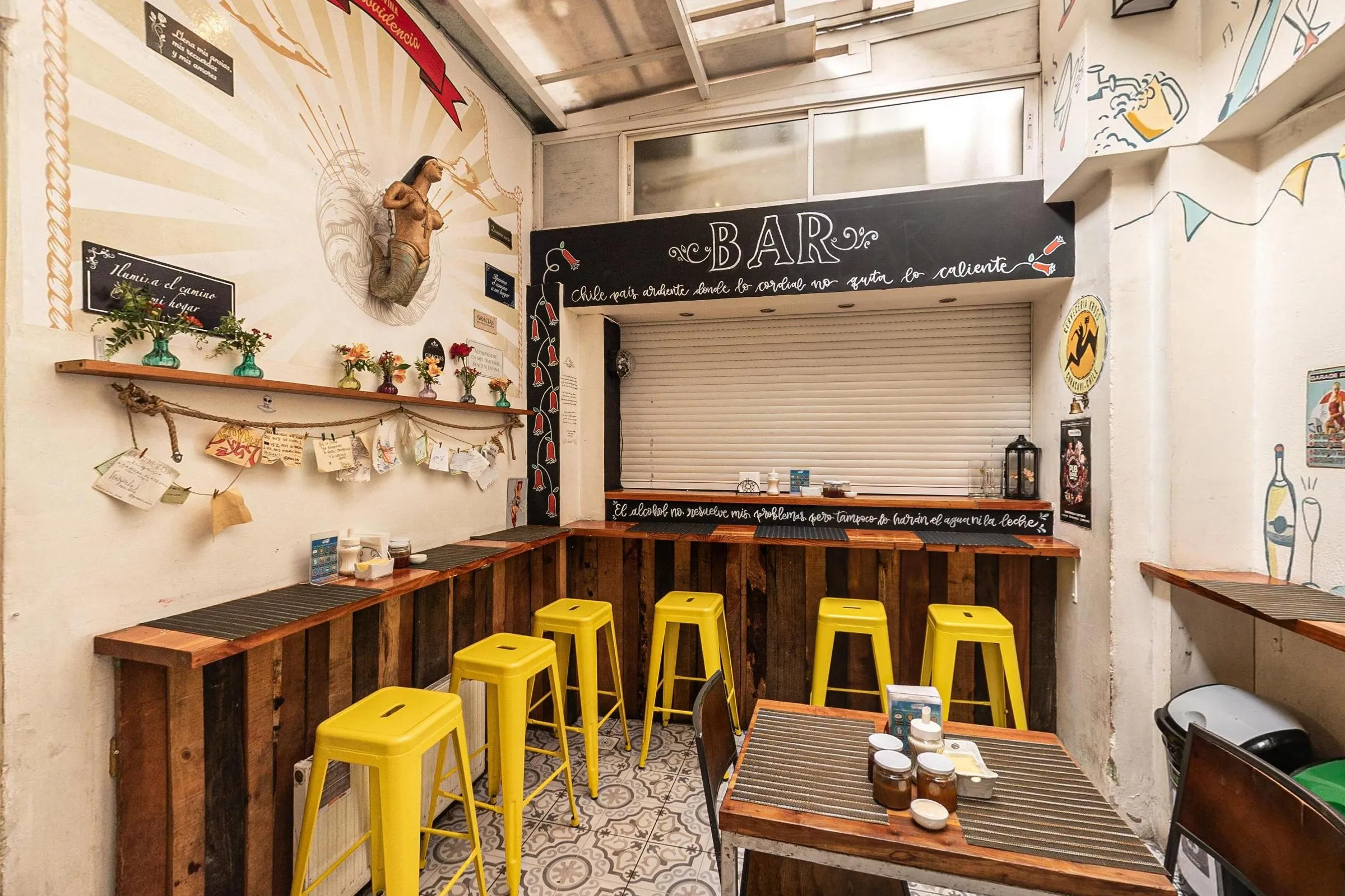 Bar com balcão de tábuas de madeira, cinco banquetas metálicas amarelas, faixa preta com "BAR" e frases escritas em giz, porta de enrolar fechada, carranca de sereia na parede e prateleira com vasinhos de flores
