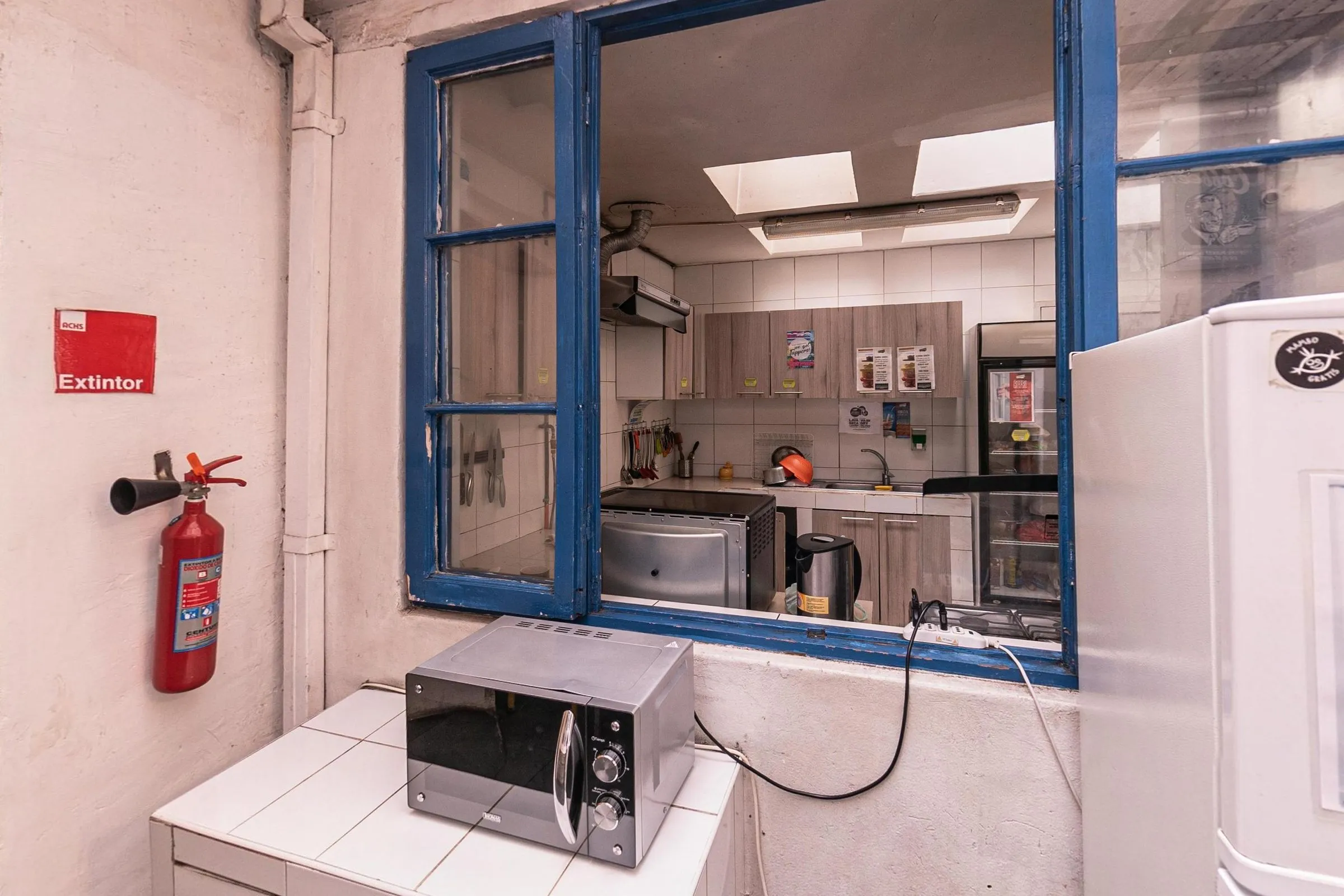 Cozinha compartilhada vista por uma janela larga de caixilho azul, com armários, micro-ondas, fogão e cafeteira; na frente, outro micro-ondas sobre a bancada de azulejo branco, um extintor e a geladeira à direita