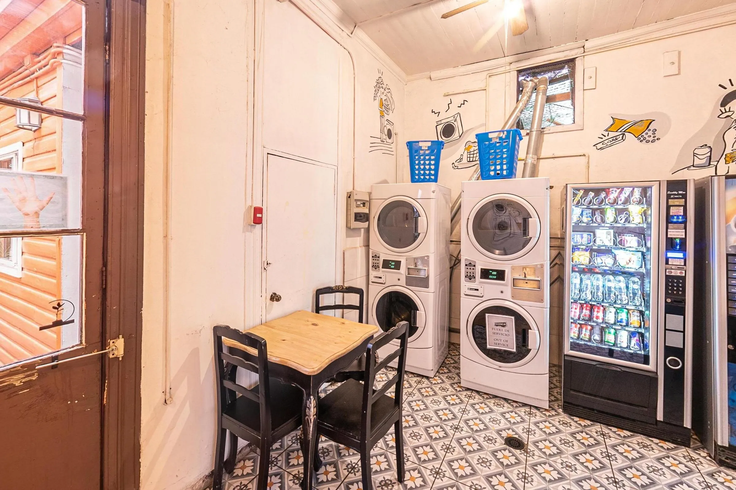 Lavanderia com duas torres de lavadora e secadora, cestos azuis, máquina de lanches e de bebidas e uma mesa de madeira clara com cadeiras pretas sobre piso de ladrilho hidráulico; paredes com desenhos em traço preto