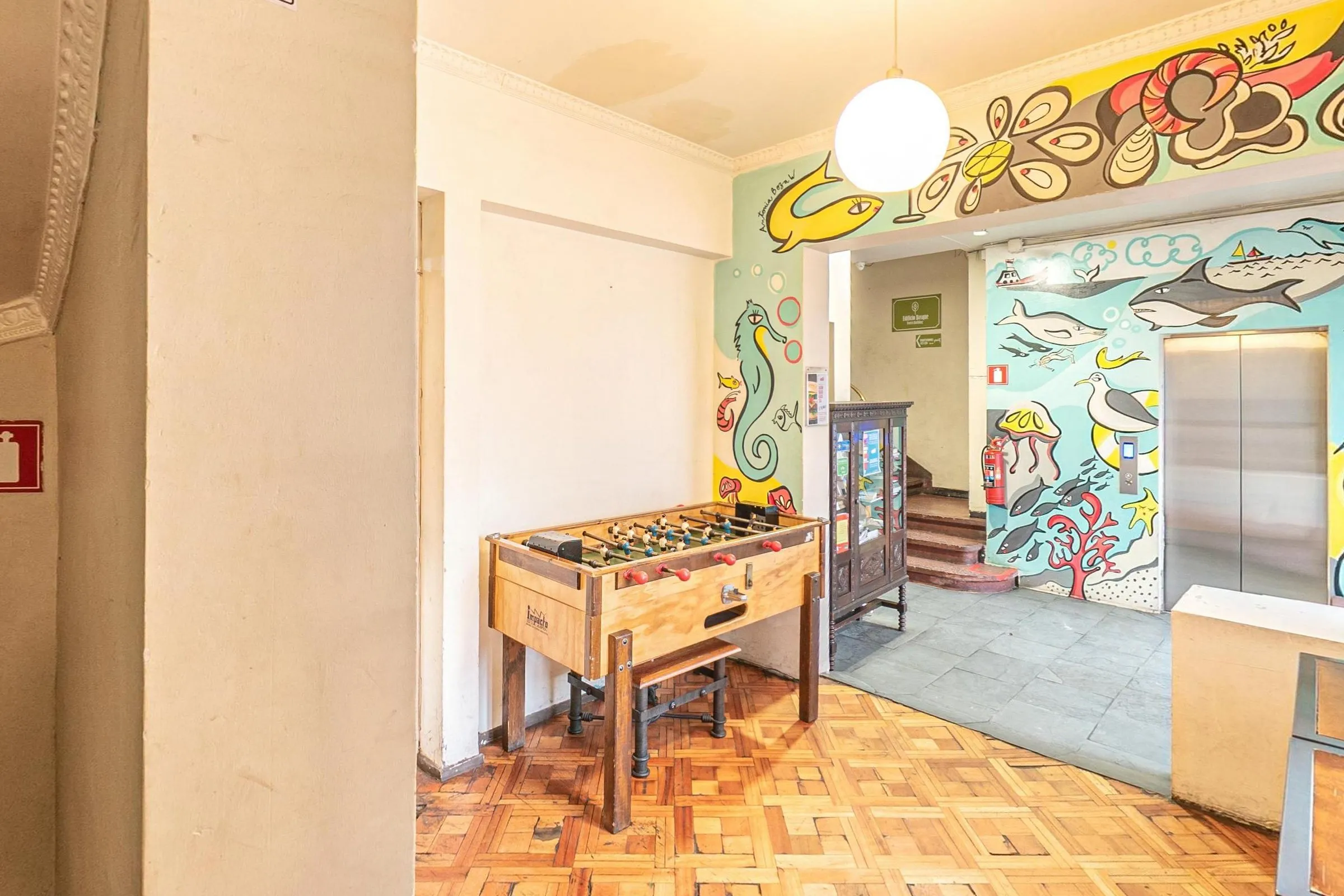 Mesa de pebolim de madeira sobre piso de taco, com o hall do elevador ao fundo, paredes pintadas com mural de peixes, golfinhos e cavalos-marinhos e um lustre de globo branco