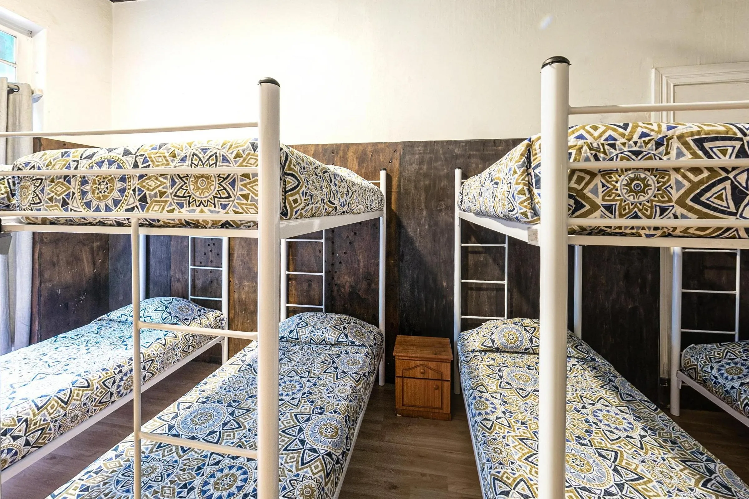 Dormitório de 8 camas do Hostal Providencia com beliches de metal branco e roupa de cama estampada em azul, amarelo e branco, painel de madeira escura na parede, piso de madeira clara e um baú entre as camas