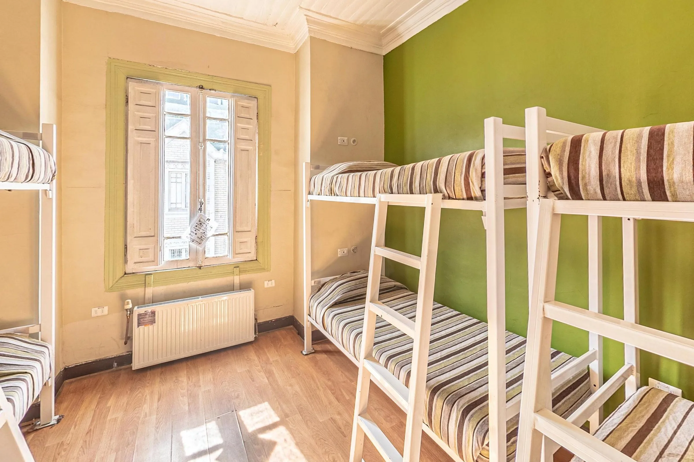 Dormitório de 6 camas com beliches brancos de escada inclinada, colchas listradas em marrom e bege, parede verde-oliva, janela de venezianas de madeira com moldura verde e aquecedor abaixo