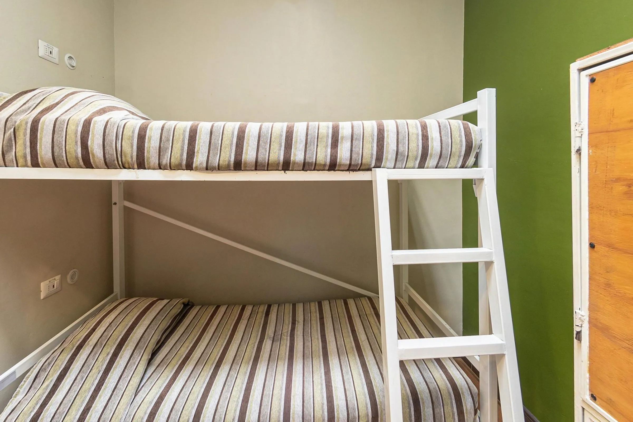 Beliche branco com colchas listradas em marrom, bege e branco no dormitório de 4 camas do Hostal Providencia, entre uma parede cinza-clara e outra verde, com um armário de compensado à direita