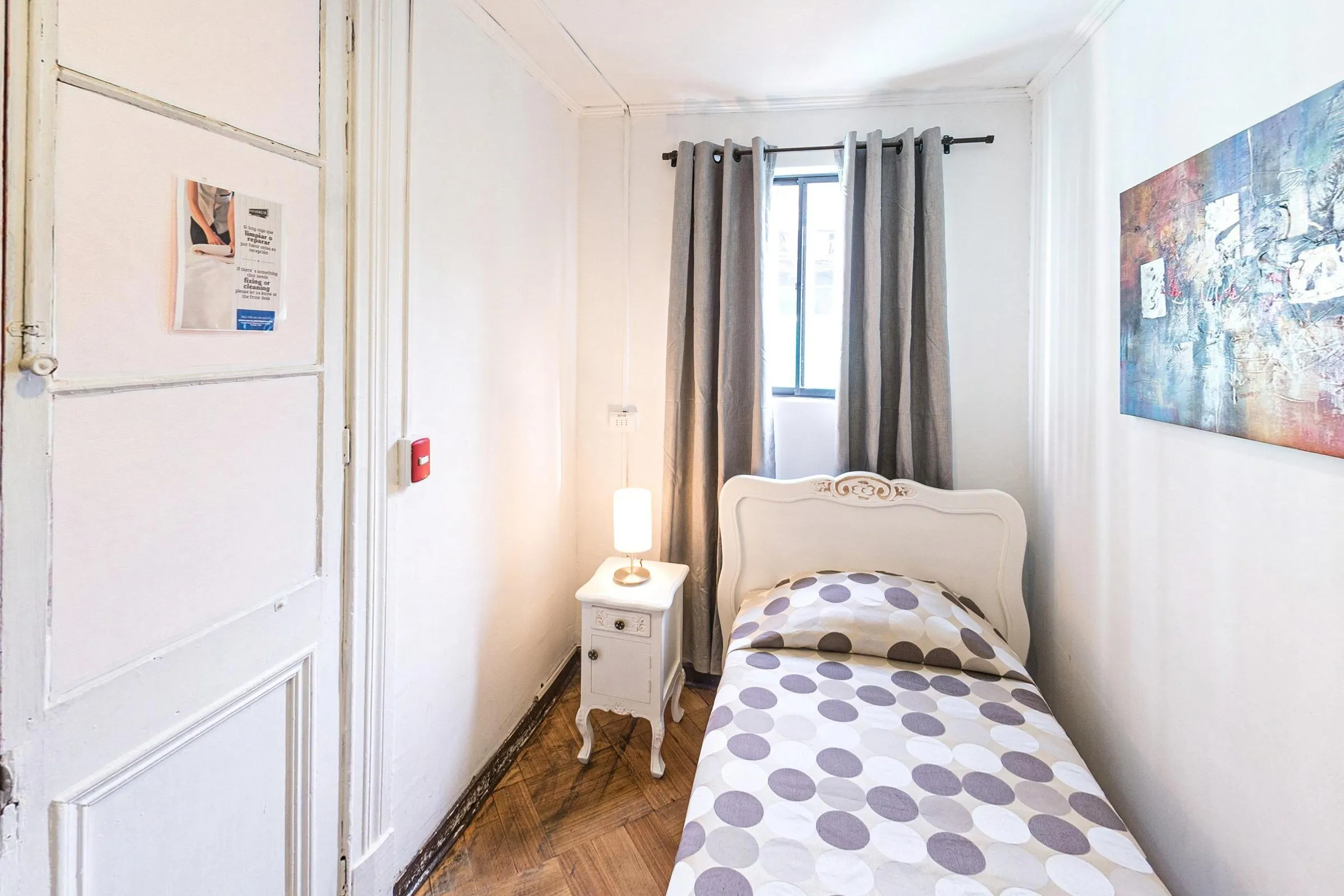 Quarto individual com cama de solteiro de cabeceira branca entalhada, colcha de bolinhas cinza, criado-mudo branco com abajur, cortinas cinza na janela e um quadro abstrato na parede