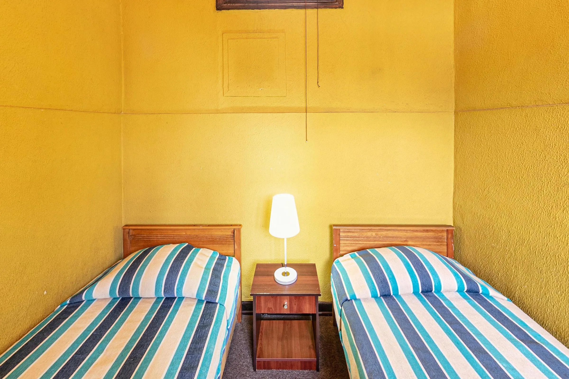 Quarto twin com duas camas de solteiro de cabeceira de madeira e colchas listradas em azul, branco e verde-água, criado-mudo com abajur entre elas e paredes amarelas