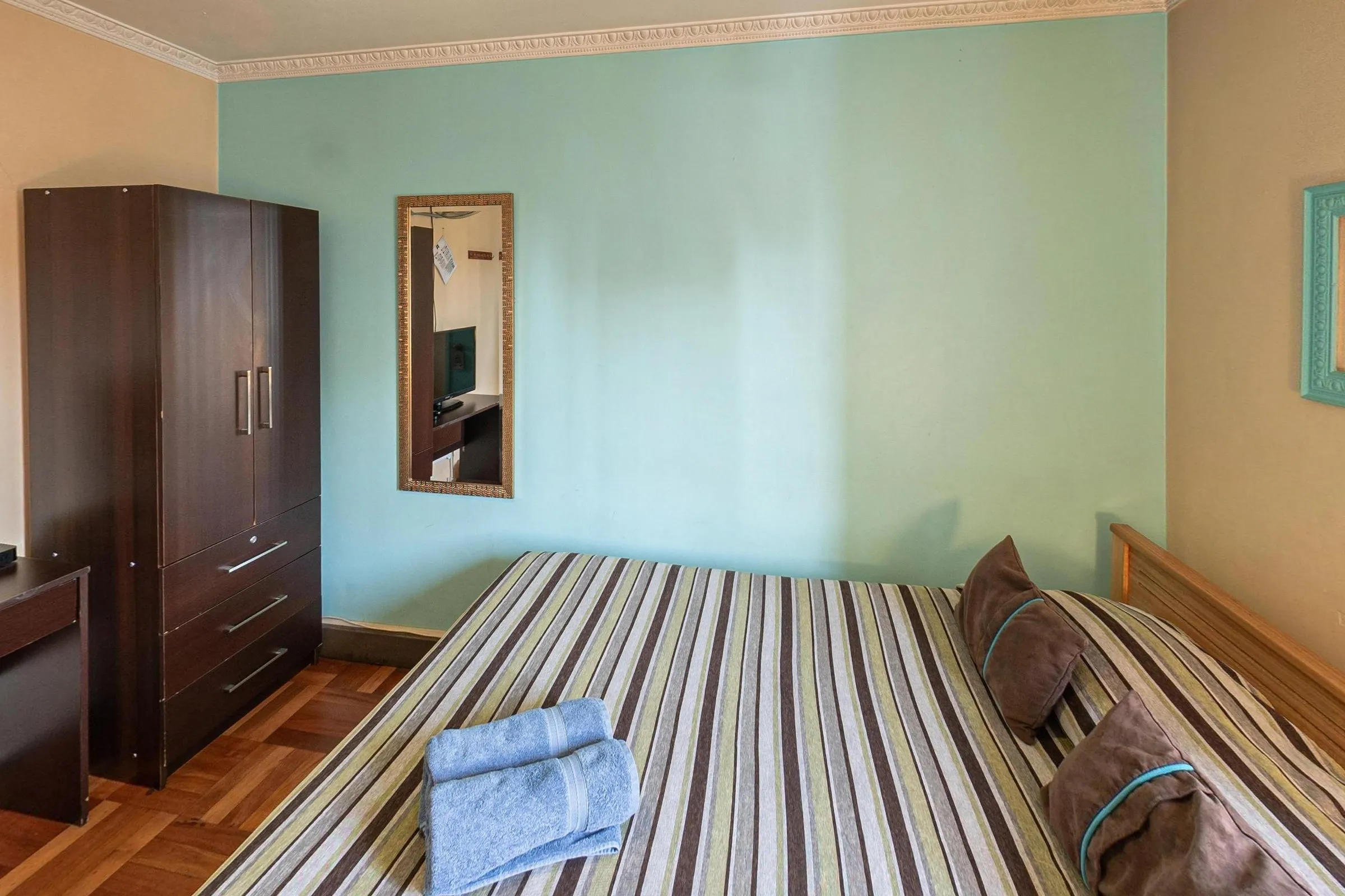 Quarto de casal com cama de casal de colcha listrada, almofadas marrons e toalhas azuis, armário de madeira escura, espelho de moldura dourada e parede verde-água