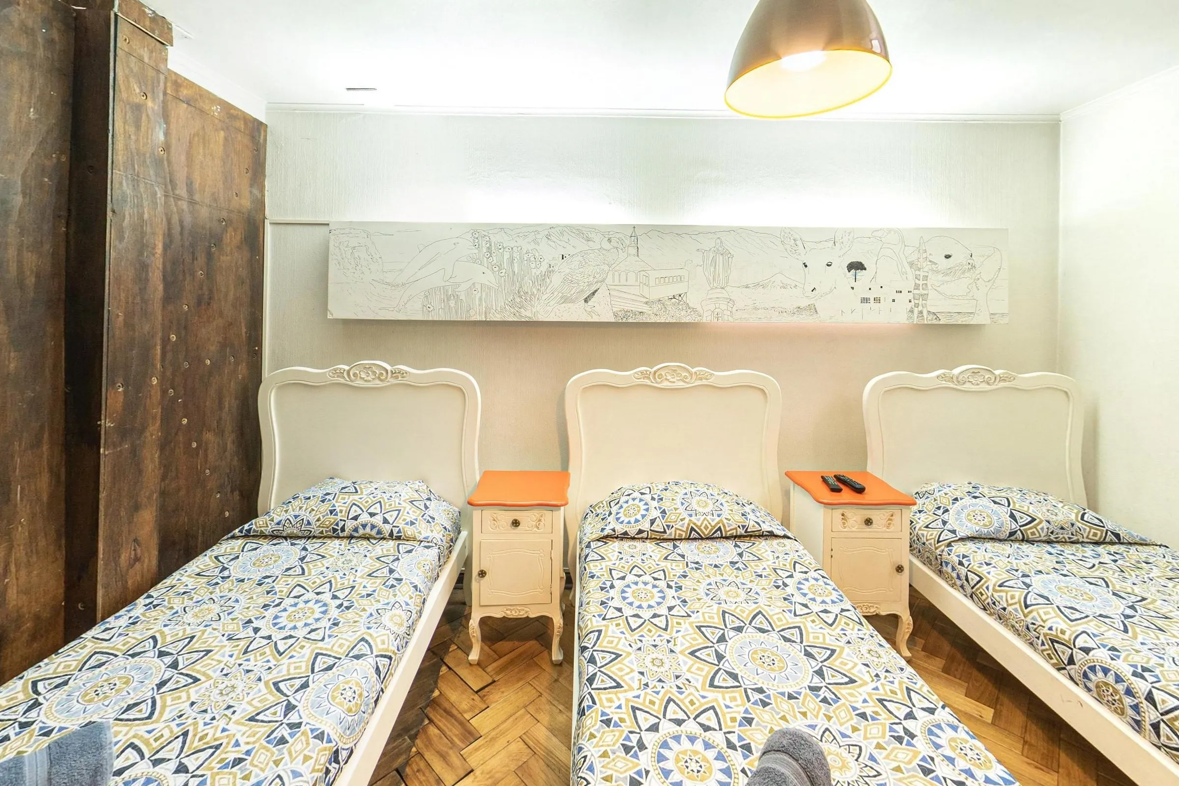 Quarto triplo com três camas de solteiro de cabeceira branca entalhada lado a lado, colchas estampadas em azul e amarelo, dois criados-mudos com tampo laranja, painel desenhado em preto e branco na parede e piso de taco em espinha