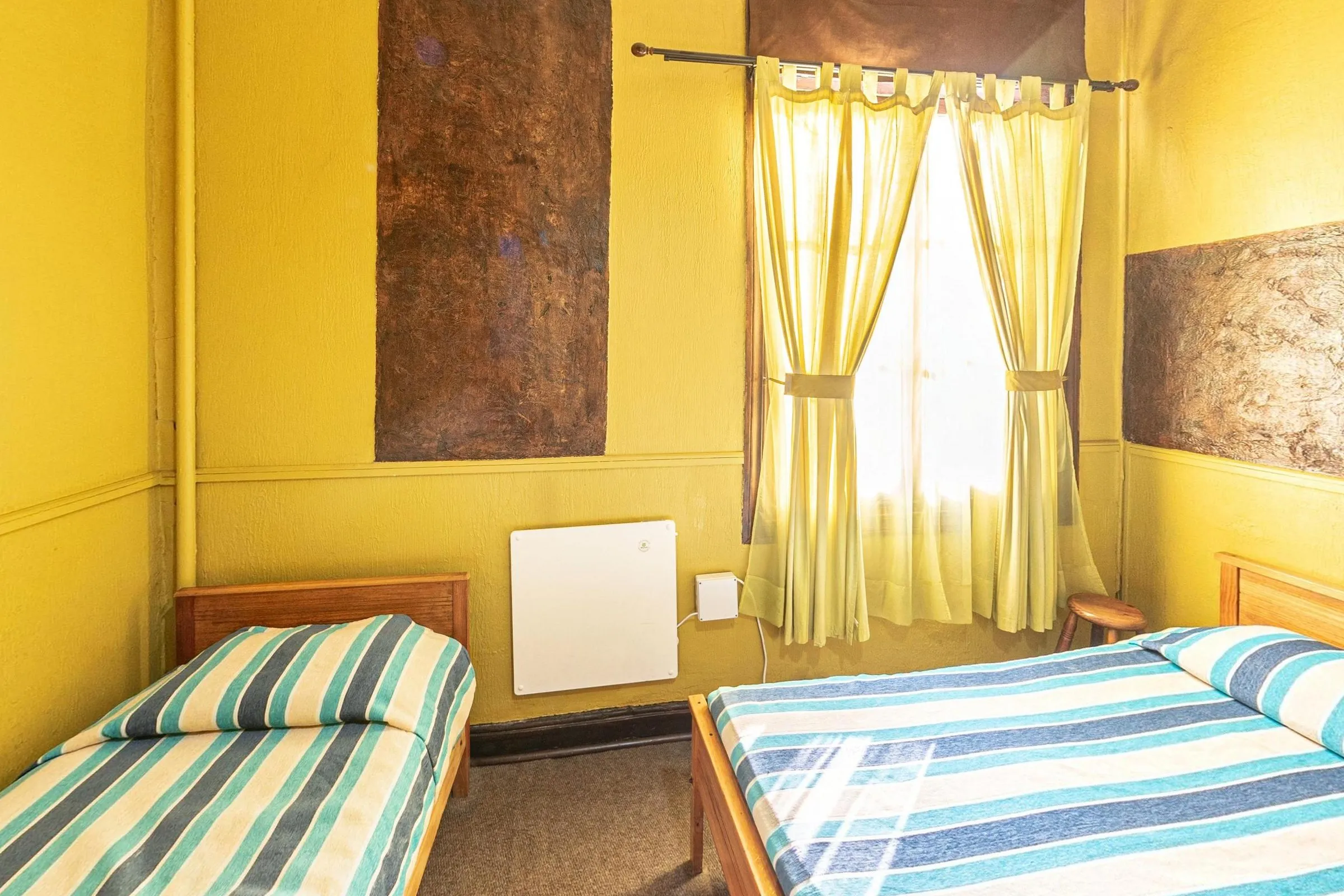 Quarto triplo com banheiro privativo, com uma cama de solteiro e uma de casal de colchas listradas em azul e verde-água, paredes amarelas com painéis de textura marrom, janela com cortinas amarelas e aquecedor branco
