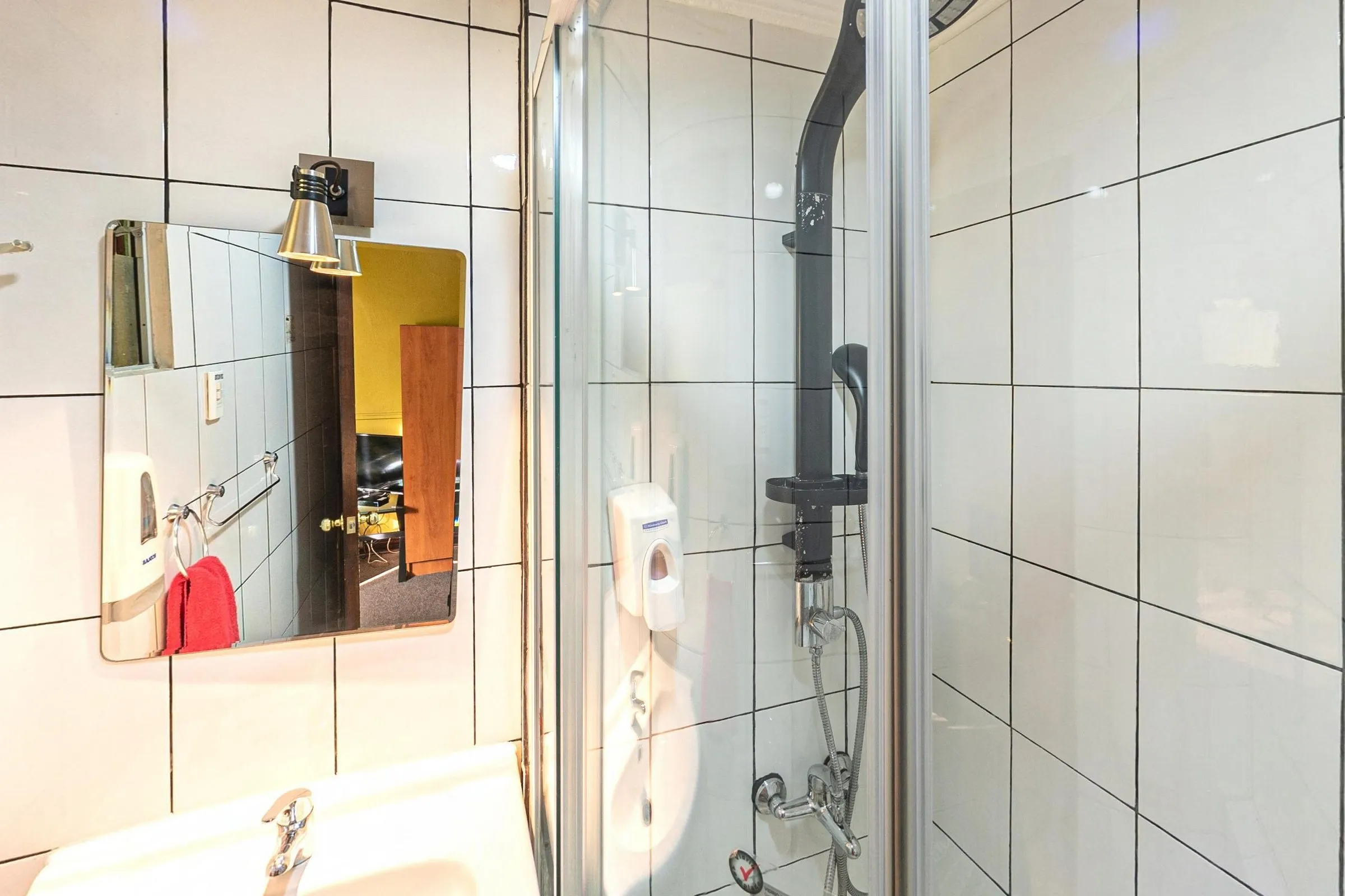 Banheiro privativo do quarto triplo do Hostal Providencia, com azulejos brancos, pia com espelho e arandela, toalha vermelha, dispensers de sabonete e box de vidro com chuveiro preto