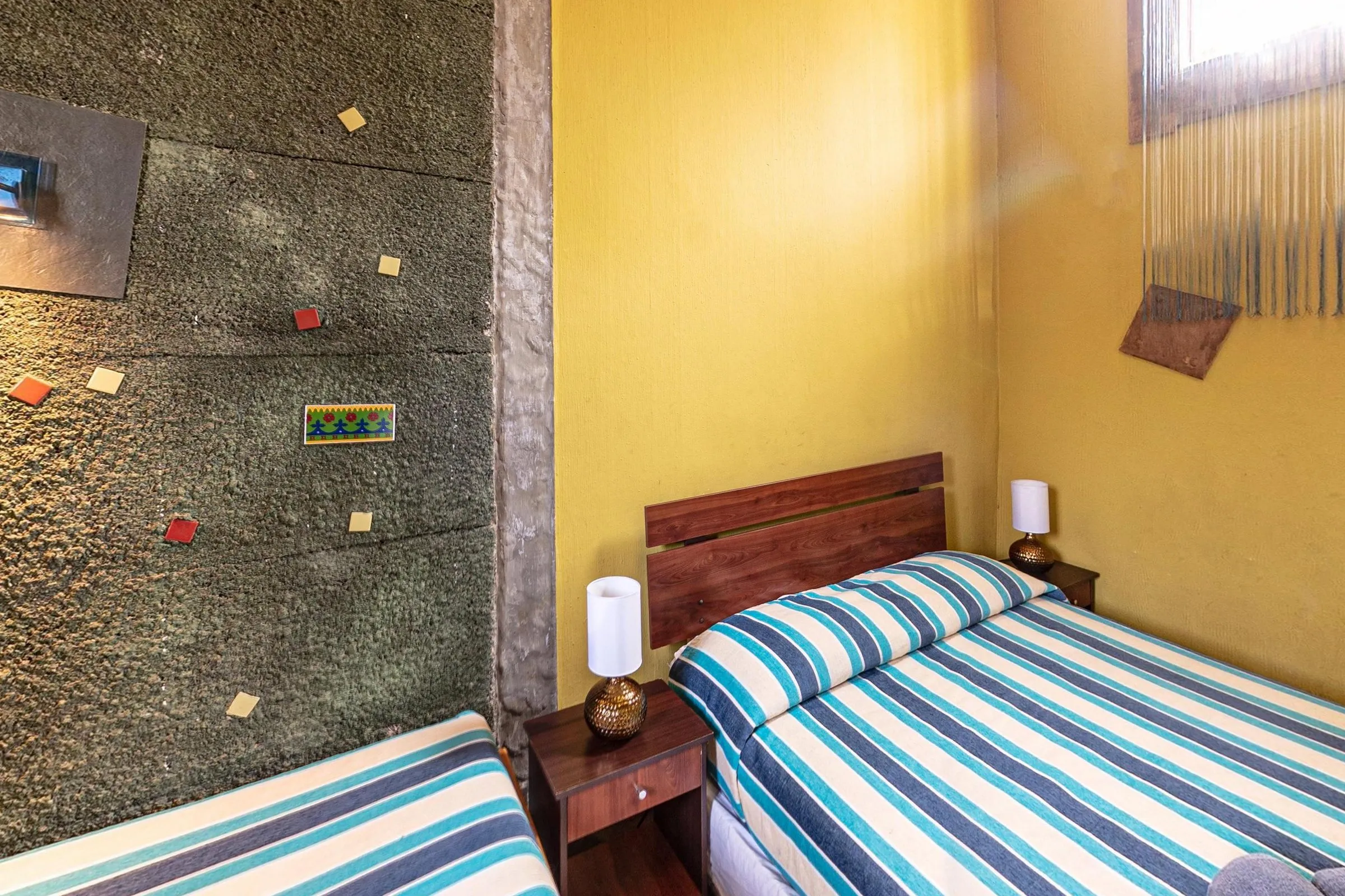 Quarto quádruplo com banheiro privativo, com cama de casal de cabeceira de madeira e uma cama de solteiro, colchas listradas em azul, branco e verde-água, parede de reboco cinza com pastilhas coloridas e paredes amarelas