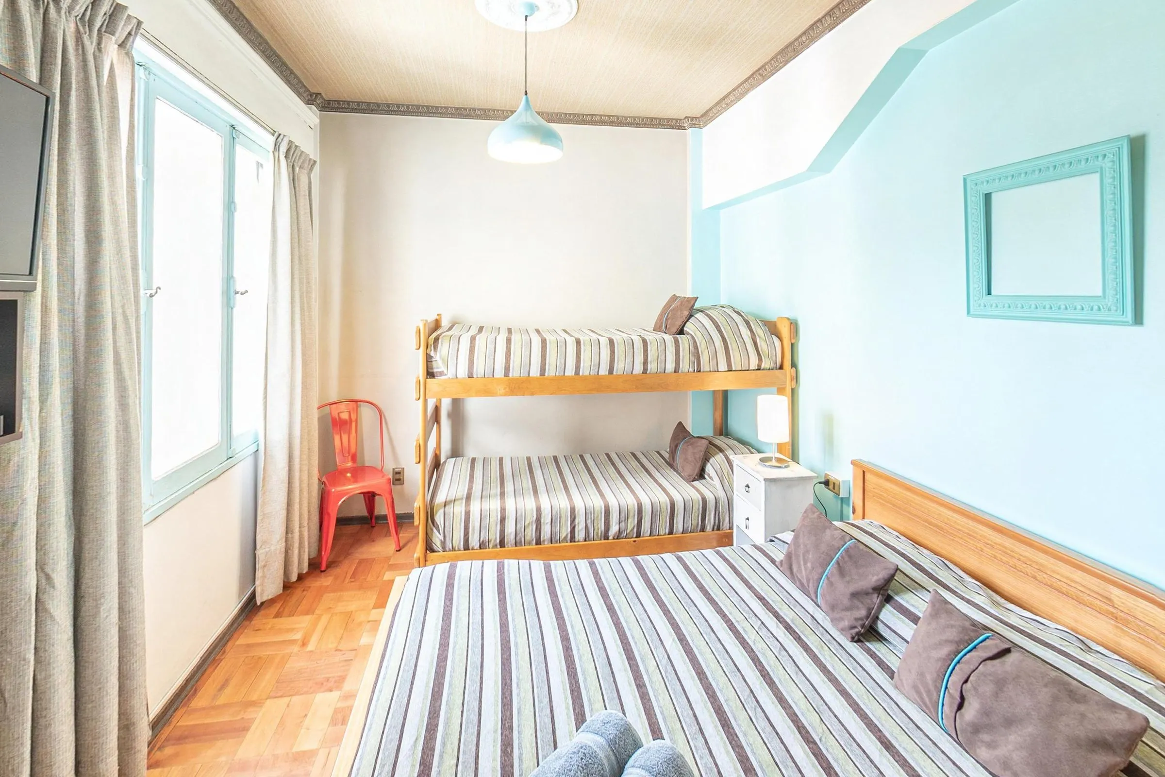 Quarto familiar do Hostal Providencia com cama de casal de colcha listrada e almofadas marrons, beliche de madeira clara ao fundo, cadeira vermelha, janela com cortinas bege, parede verde-água e luminária pendente