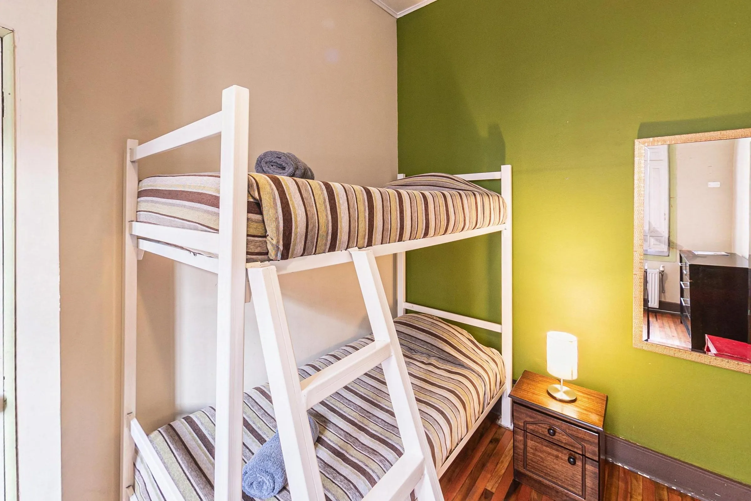 Quarto privativo com beliche branco de colchas listradas, parede verde-oliva, criado-mudo de madeira com abajur, espelho de moldura clara e piso de madeira