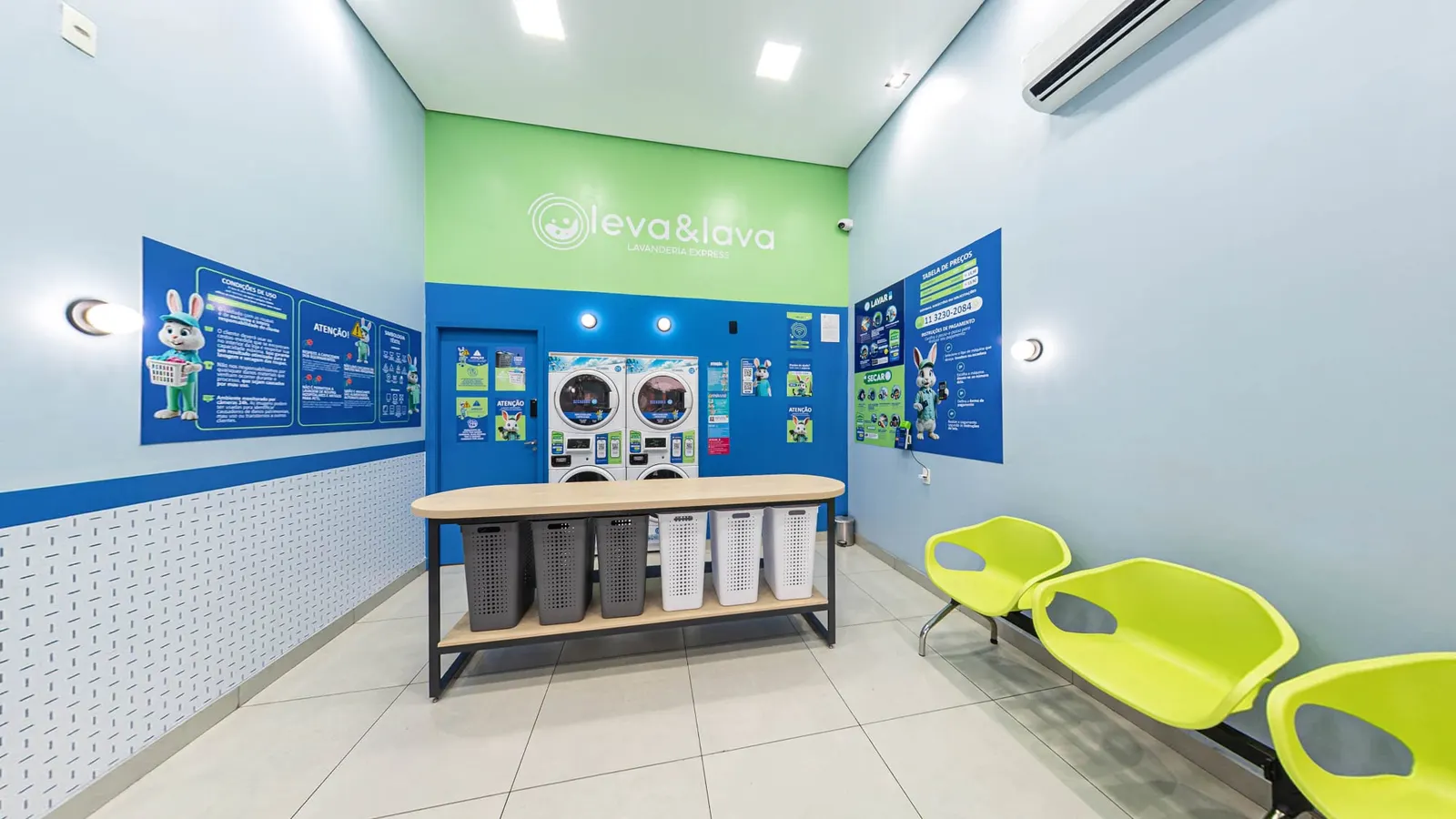 Interior da lavanderia self-service Leva & Lava, com a mesa de dobra e os cestos cinza e brancos, as secadoras empilhadas sobre as lavadoras ao fundo, cartazes de instrução nas duas paredes e cadeiras verdes de espera