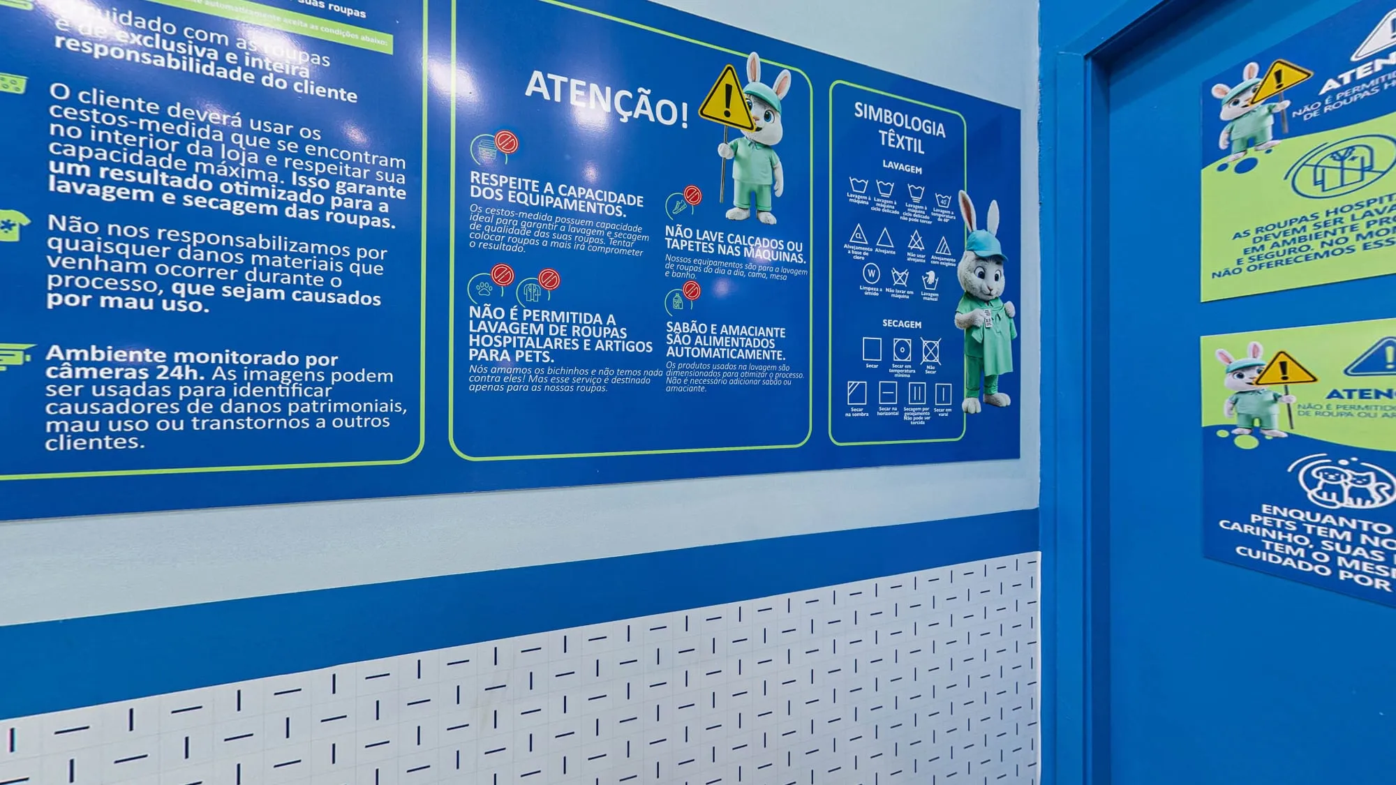 Painel azul com as condições de uso da lavanderia, os avisos de capacidade, de itens proibidos e de sabão automático e a simbologia têxtil, com o coelho mascote