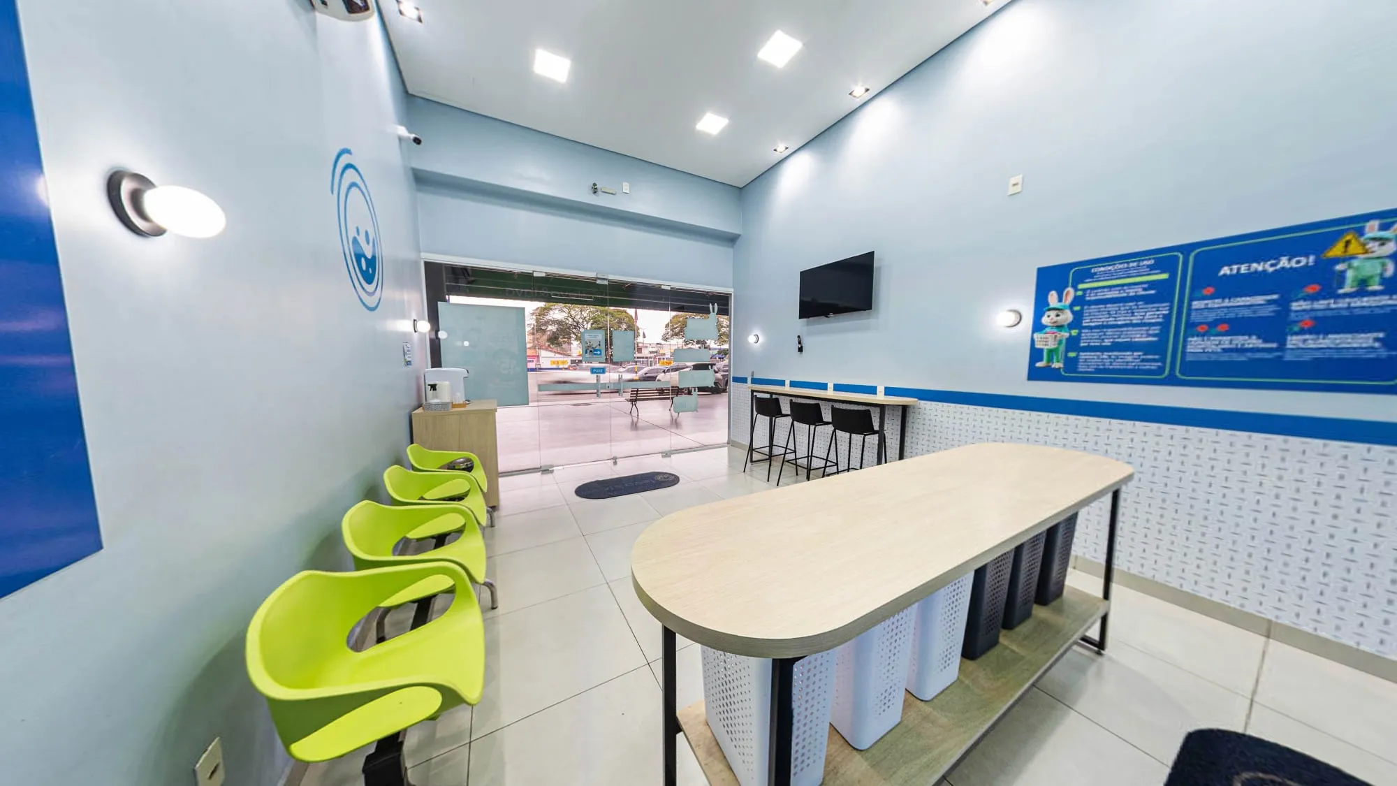 Salão da lavanderia com mesa de madeira clara, cestos embaixo, cadeiras verde-limão, balcão com banquetas pretas e a porta de vidro para a galeria